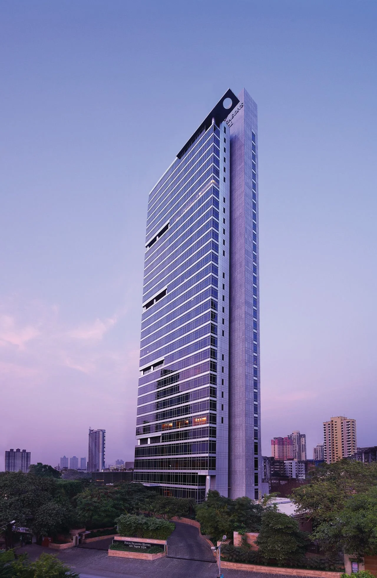 A tall modern skyscraper with glass windows and a sleek design, surrounded by smaller buildings in a cityscape at dusk with a purple sky.
