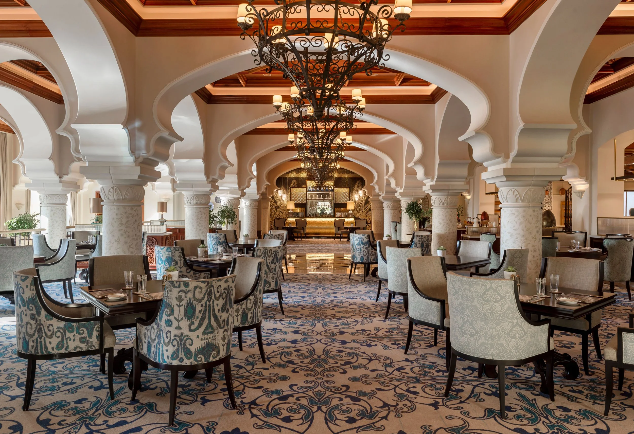 Elegant dining area with patterned upholstered chairs, round tables set with glassware, and a patterned carpet. The space features ornate white arches, columns with intricate designs, and a large central chandelier with multiple lampshades hanging from a decorated ceiling. A bar or service area is visible in the background, and the room is decorated with plants and lamps, creating a luxurious atmosphere.