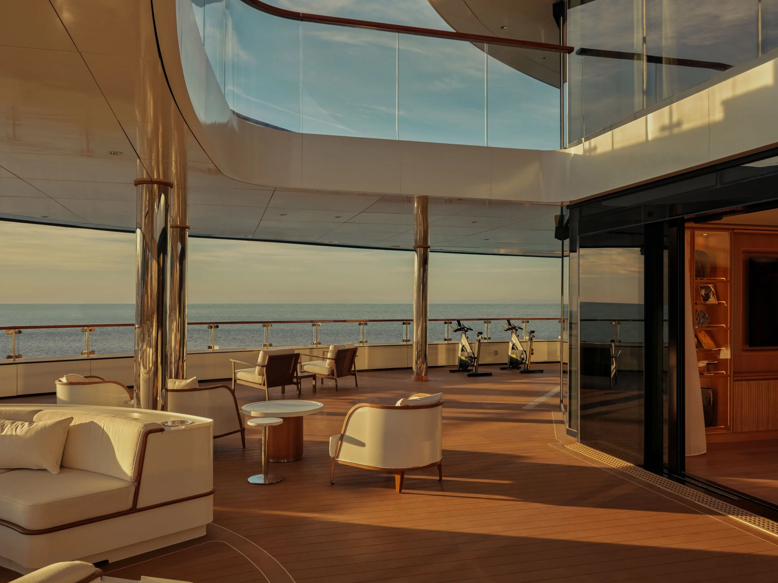 A luxurious cruise ship deck overlooking the ocean at sunset, with modern white chairs, small tables, and bicycles, enclosed partially by glass walls and supported by polished metal columns.