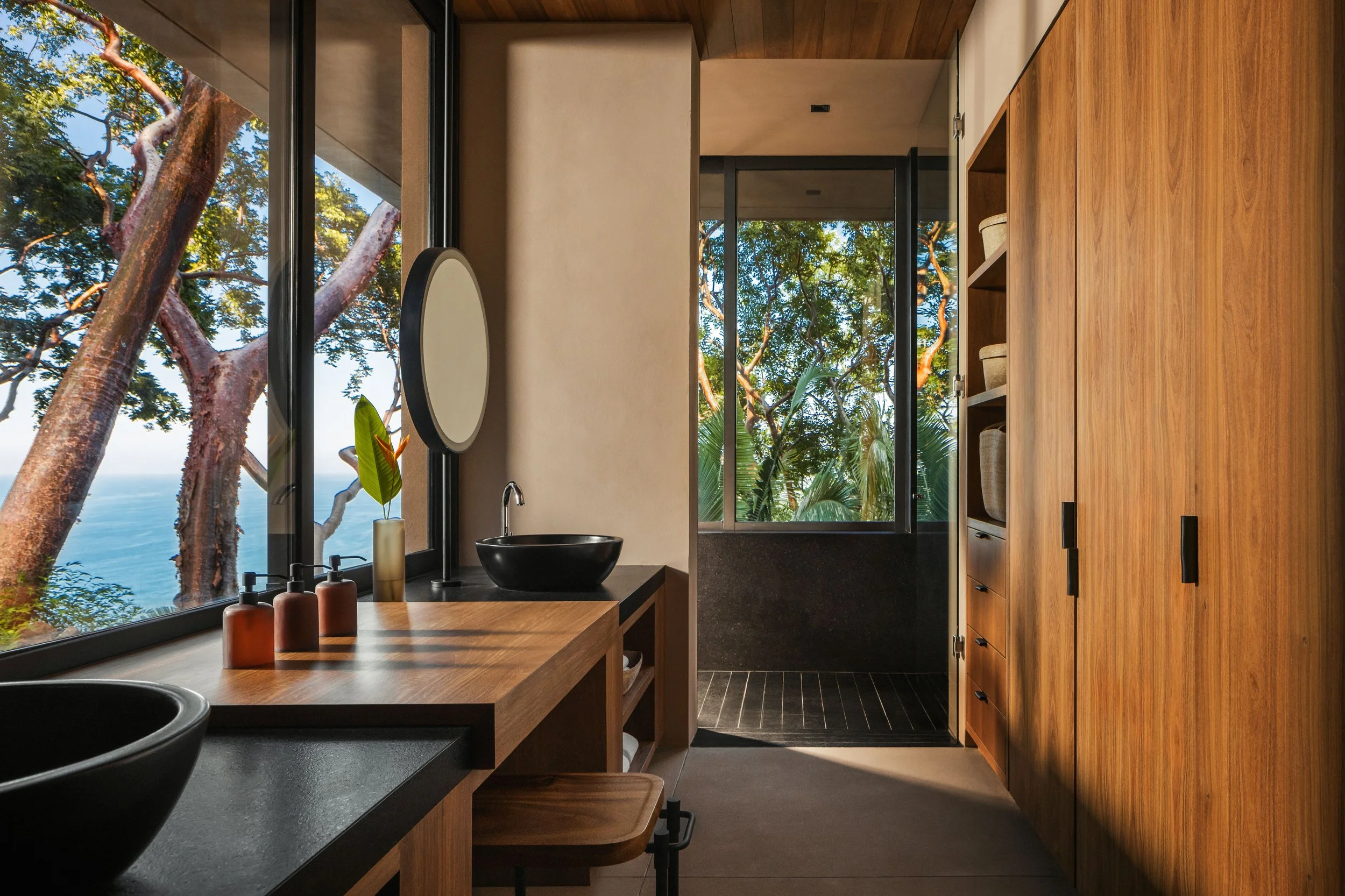 Contemporary bathroom with wooden cabinets and large windows showing trees outside. A black vessel sink, a mirror, and a few soap dispensers are on the countertop.