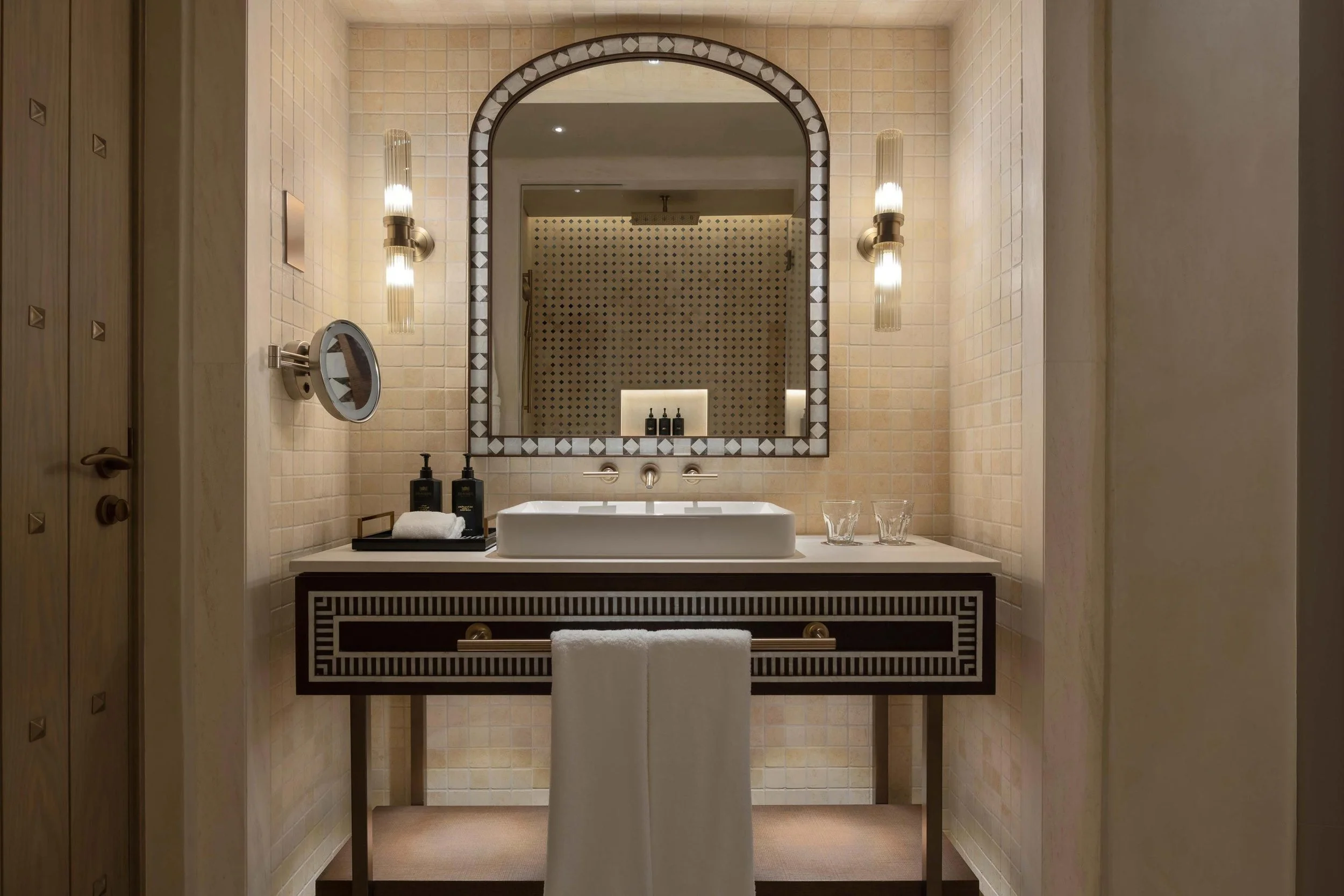 A modern bathroom vanity with a rectangular vessel sink, black and white decorative border mirror, two gold and glass wall sconces, black soap dispensers, glasses, white towel on a black stand, and beige tiled walls.