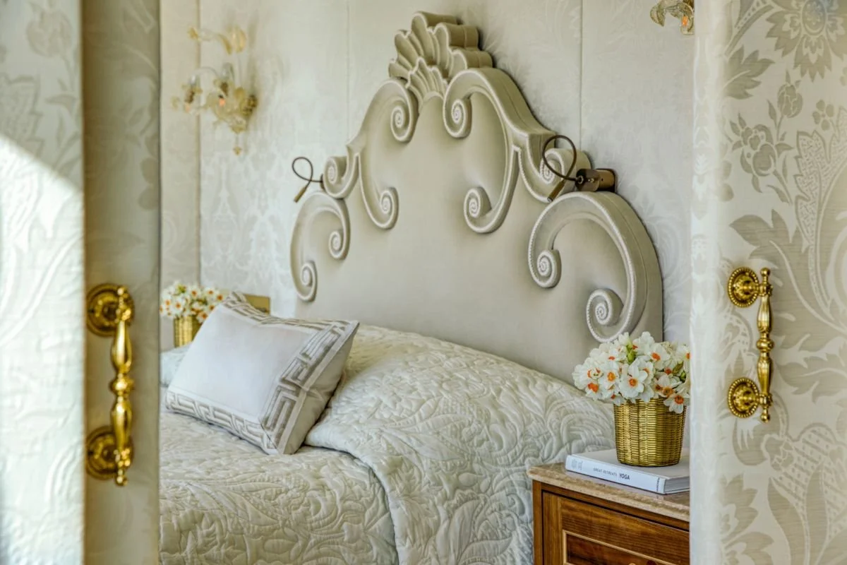 A luxurious bedroom featuring an ornate, cream-colored headboard with scroll and floral designs, a bed with a textured cream bedspread and matching pillow. There is a wooden nightstand with a wicker basket filled with white and orange flowers and a couple of books, one titled 'Yoga'. The walls and curtains have a elegant floral pattern, complemented by golden curtain tiebacks and wall fixtures.