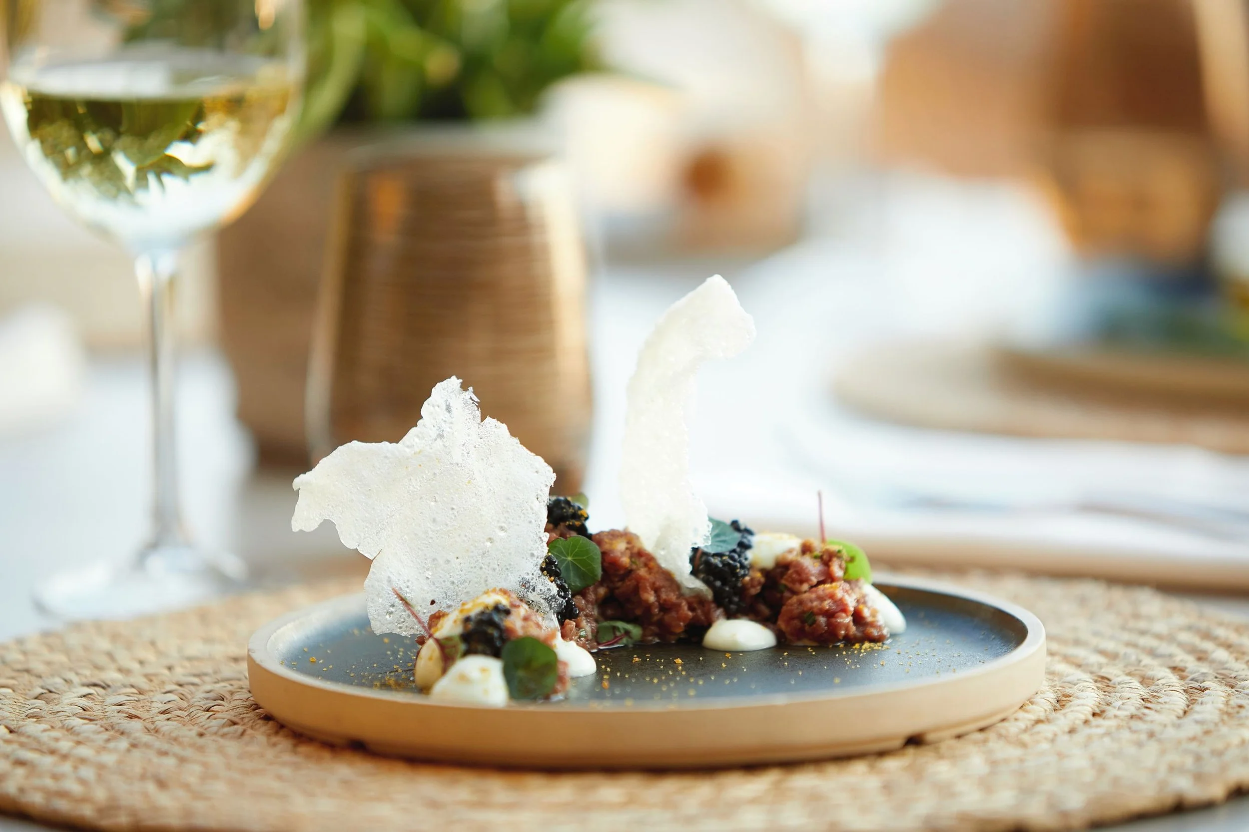 A plated dish with a white dollop and garnishes in front of a glass of white wine, with a blurred background of a table setting.
