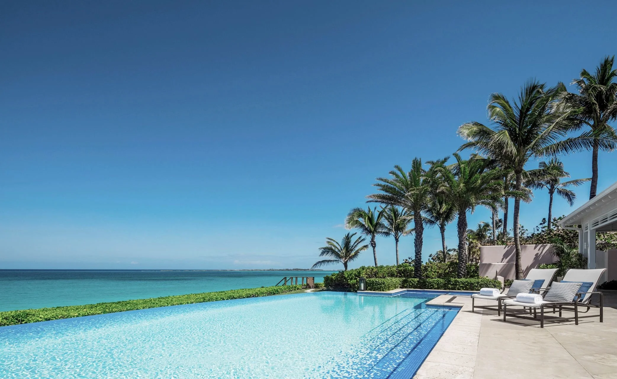 Luxury resort with a large swimming pool, palm trees, and outdoor furniture overlooking the ocean under a clear blue sky.