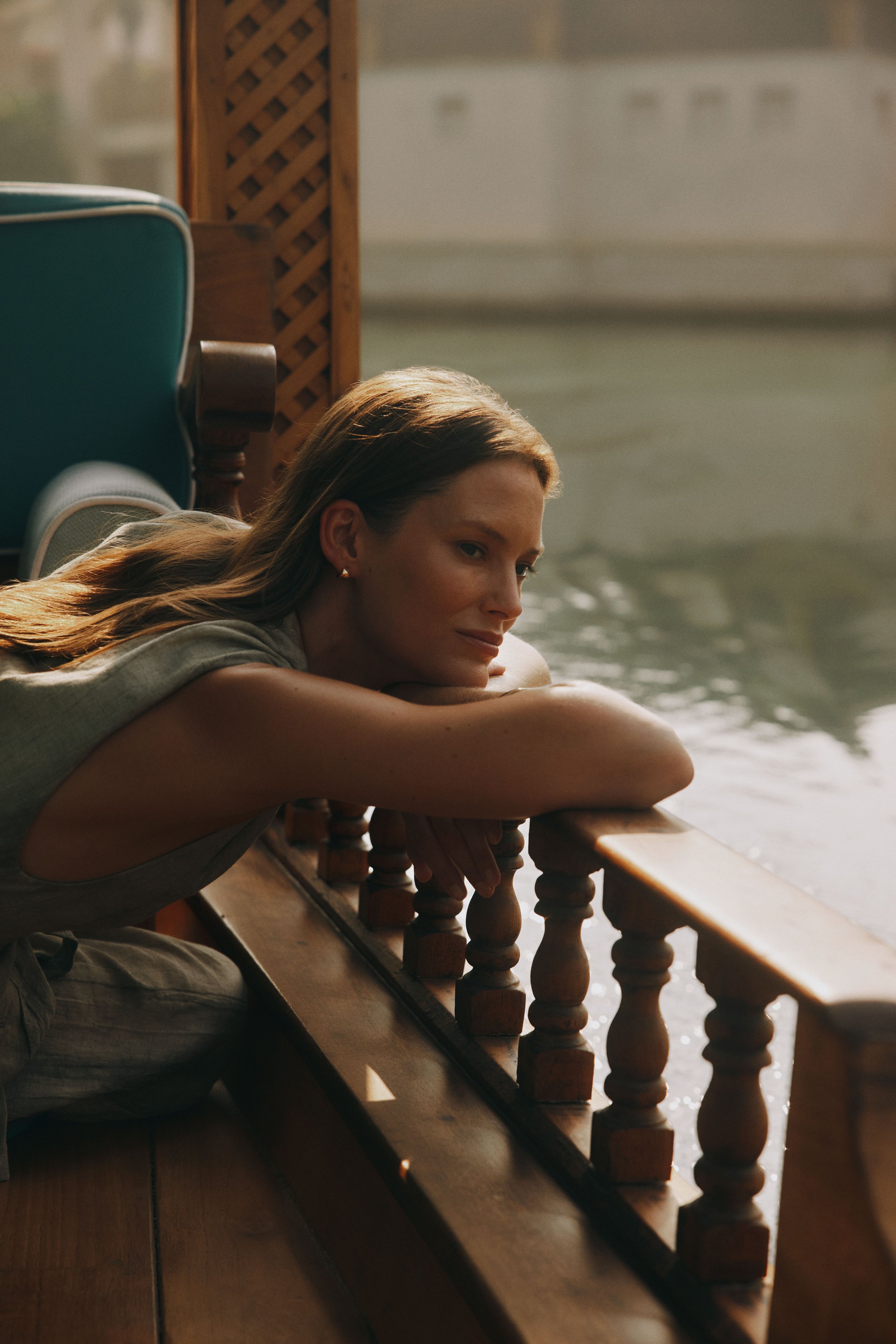 A woman with long brown hair lying on her side at a wooden railing, gazing outside near a body of water with her arms folded under her head.