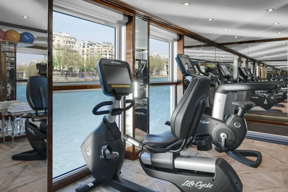Indoor gym with exercise equipment, including a stationary bike, facing large windows with a view of water and city buildings outside.