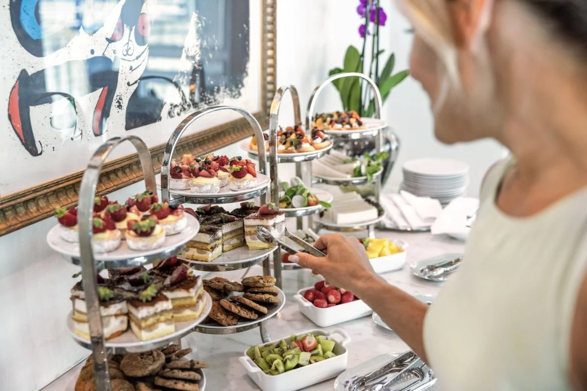 A woman selecting desserts from a multi-tiered dessert display at a buffet. The display features cakes, fruit tarts, and cookies. There are bowls of fresh fruit including strawberries, kiwi, and other fruit on the table.