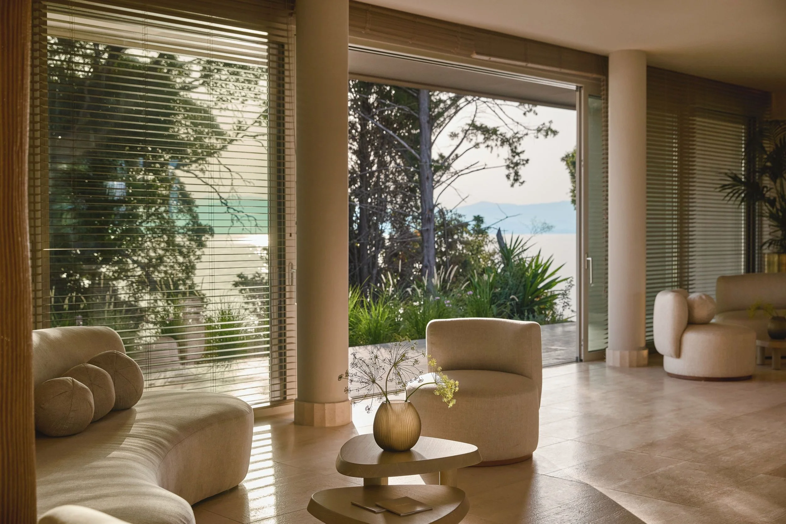 Living room with large windows, beige sofa, round coffee table with a plant, and view of trees outside.