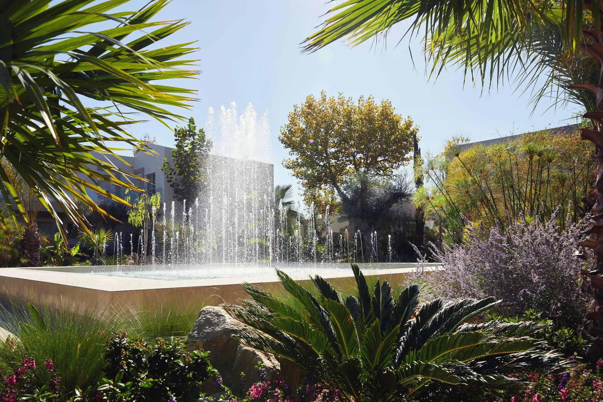 A lush garden with various green plants and trees surrounding a fountain with multiple streams of water and a building background under a clear blue sky.