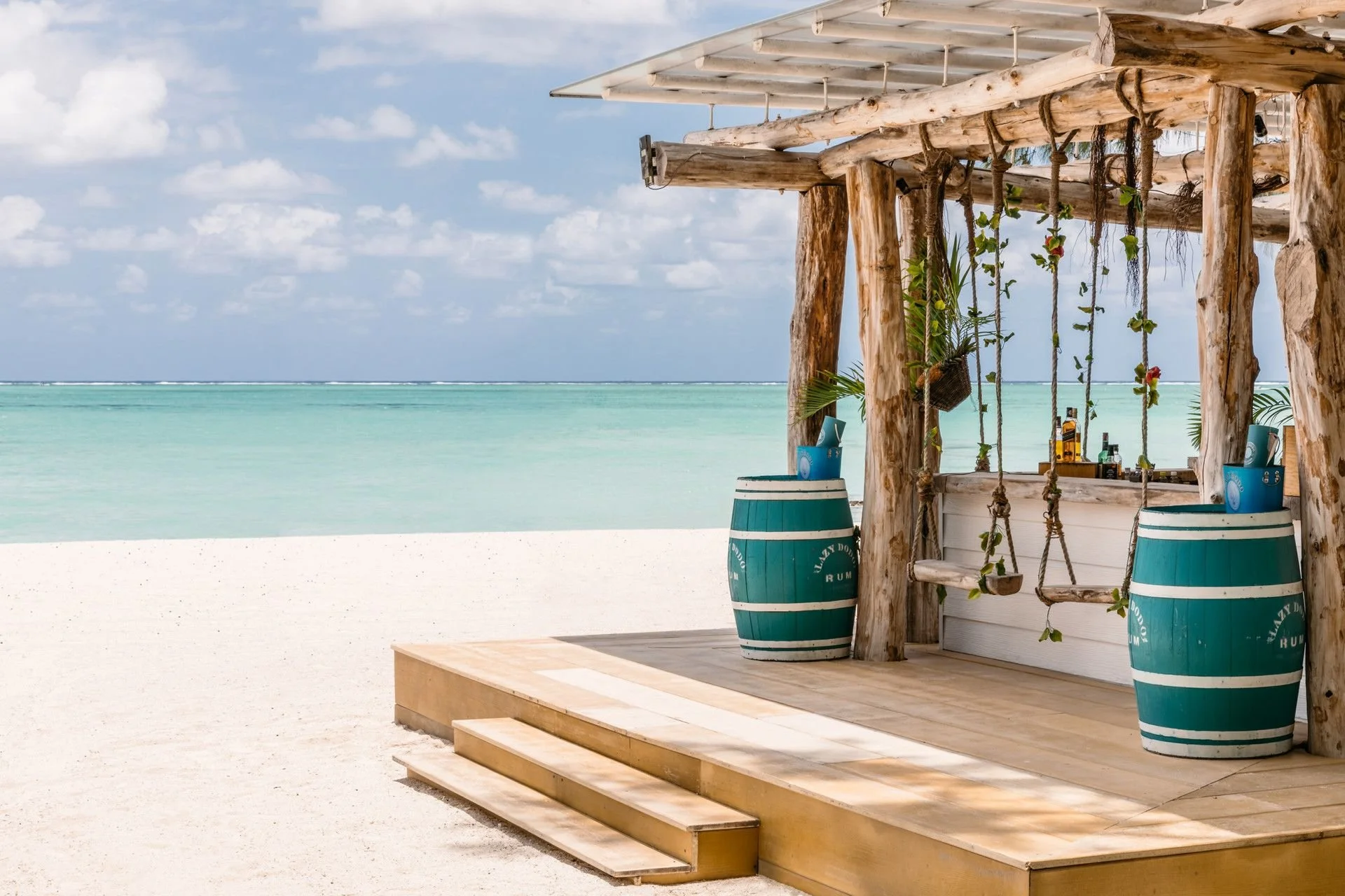 Beach bar with wooden structure, swing seats, and blue barrels, overlooking calm turquoise ocean and blue sky with scattered clouds.