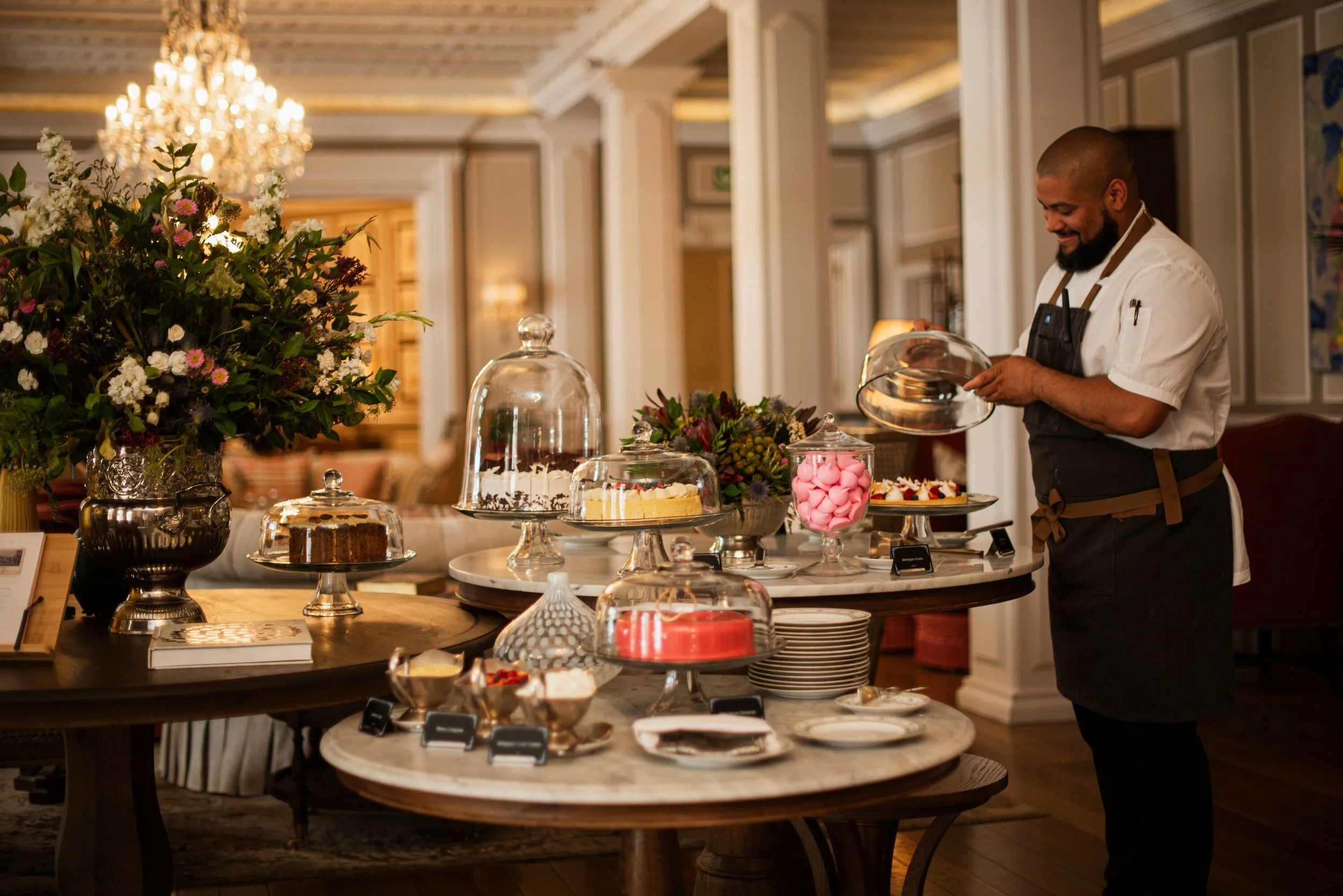 A man with a beard, wearing a white shirt and black apron, smiles as he looks at desserts on a buffet table covered with a marble top in an elegant room with a chandelier and framed art on the walls.