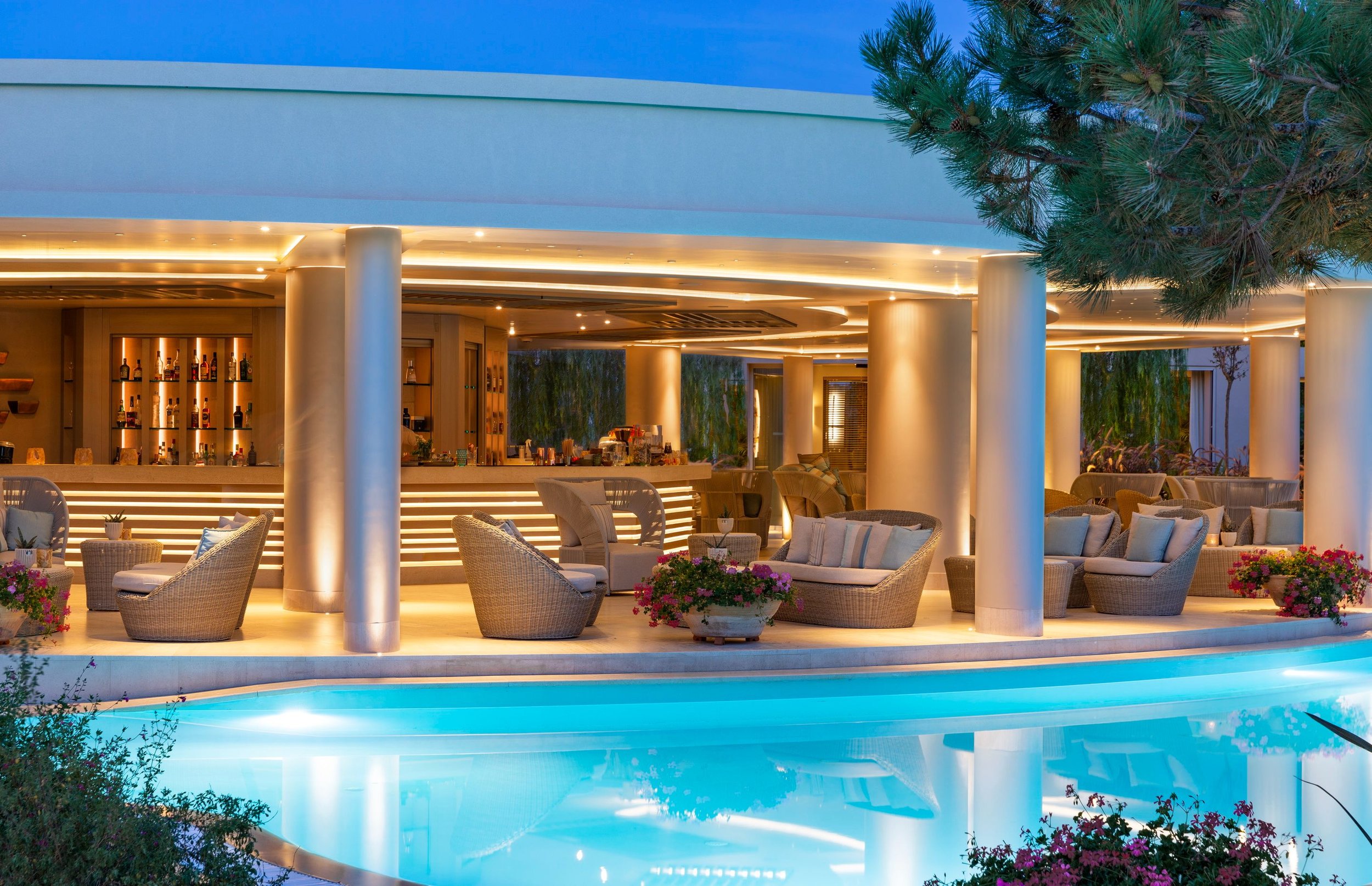 An outdoor bar and seating area next to a swimming pool, with modern lighting, wicker chairs, and potted flowers.