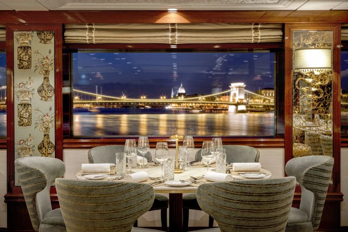 Elegant dining room set for six, with wine glasses, napkins, and plates on a round table, overlooking a cityscape with a bridge illuminated at night through a large window.