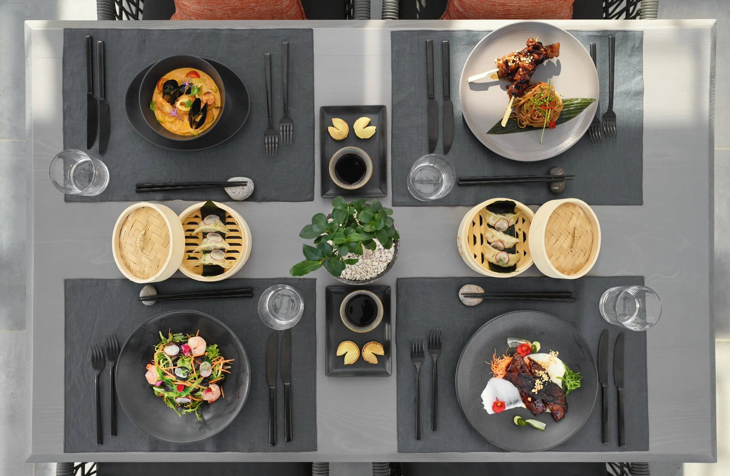 Top-down view of a dining table with an assortment of Asian dishes, including sushi, salads, noodles, and various sauces, with glasses of water and black utensils on gray placemats.