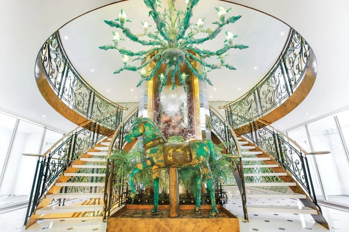 Elegant indoor space with a grand staircase, ornate wrought iron railings, a large vintage green horse statue, and a chandelier resembling a cactus with multiple arms hanging from the ceiling.
