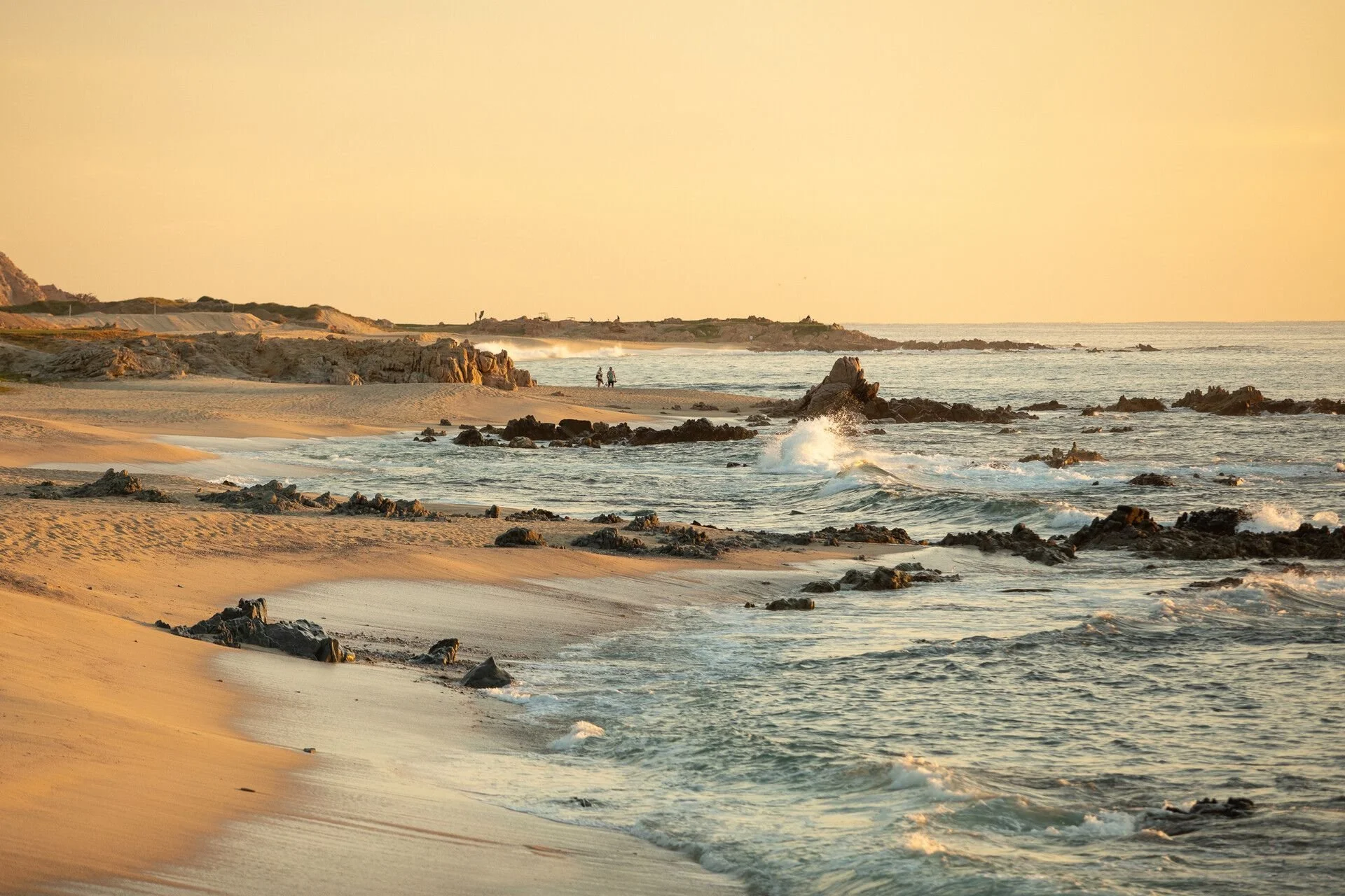 A beach scene at sunset with rocky shoreline, gentle waves, and two people walking along the shore in the distance.