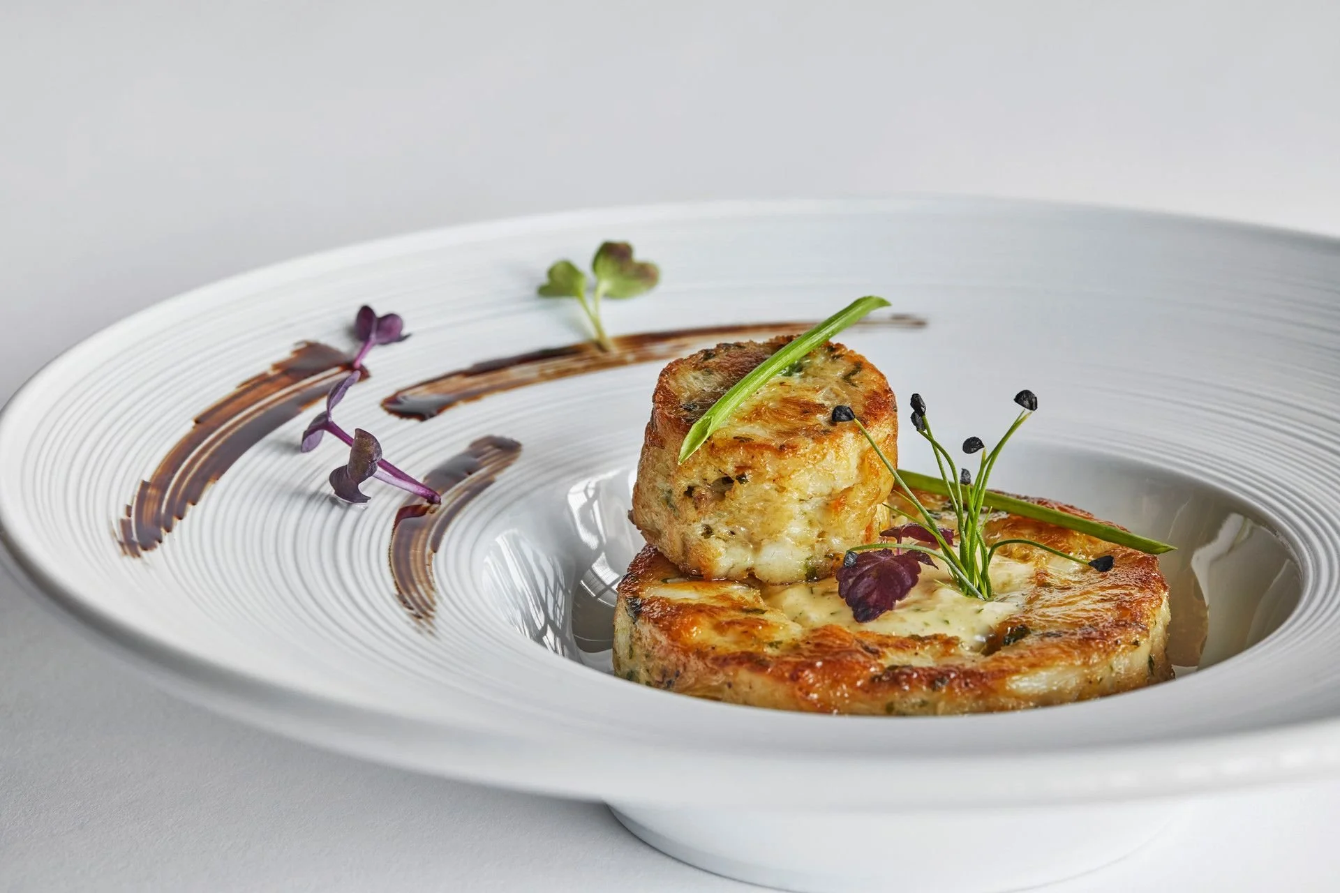Close-up of grilled crab cakes garnished with microgreens and black seeds, served on a white plate with artistic sauce decorations