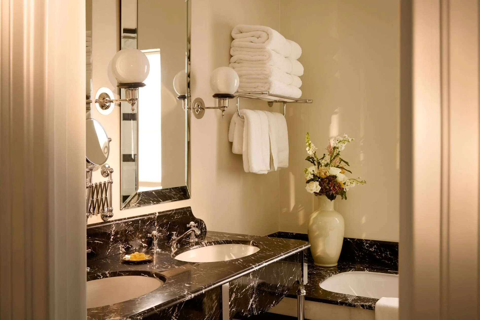 A luxurious bathroom with a black marble countertop, oval sinks, a large mirror, white towels on a metal shelf, a vase with fresh flowers, and elegant fixtures.