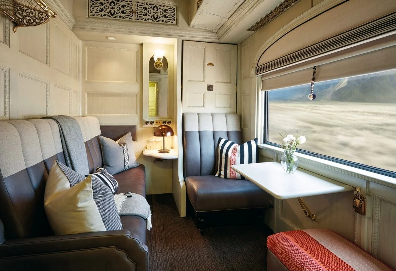 Interior of a train car with a leather couch, decorative pillows, a small side table with a lamp, and large window showing blurred landscape outside.
