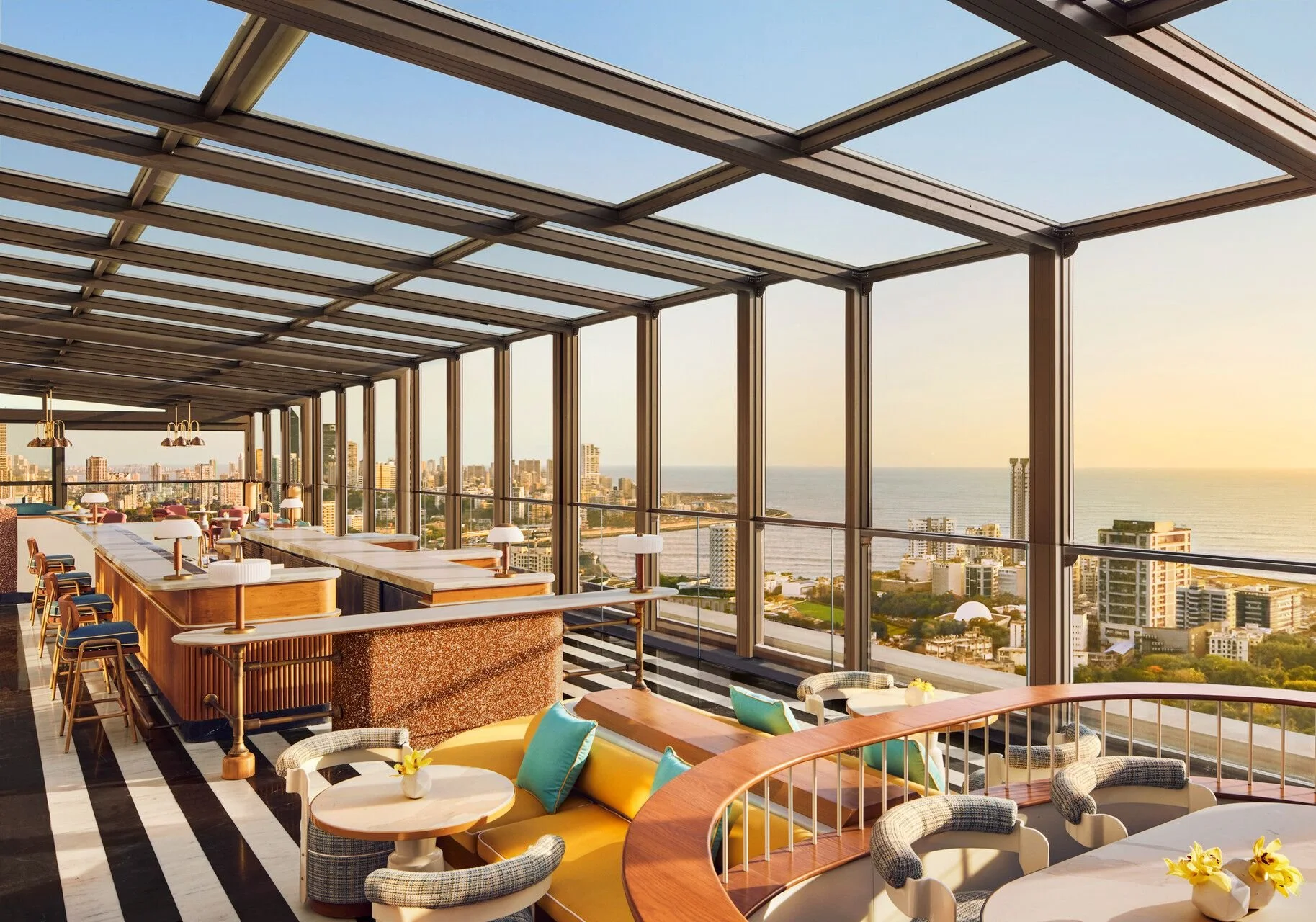 Interior of a modern high-rise restaurant or lounge with large glass windows showcasing a cityscape and ocean view, featuring stylish furniture, pendant lighting, and a black-and-white striped floor.