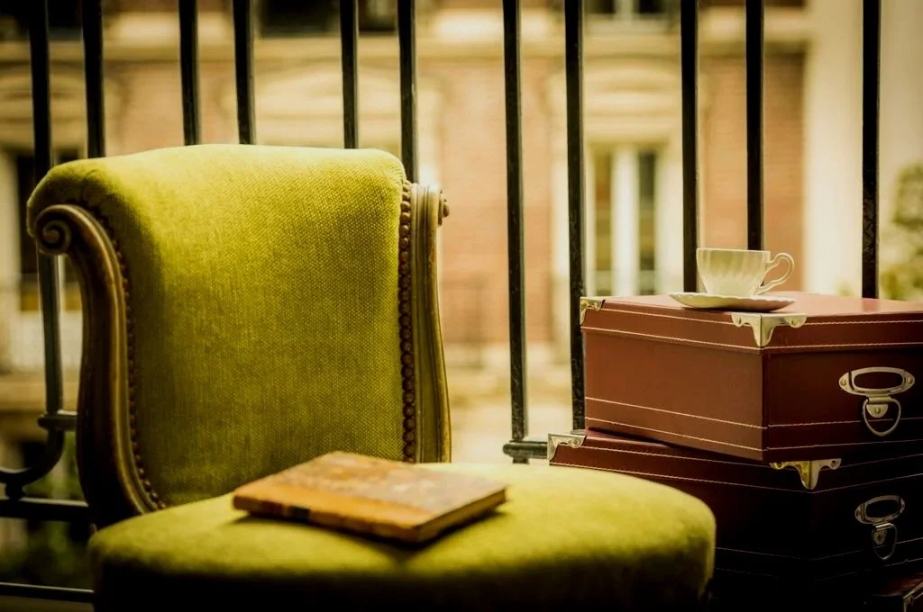Green vintage armchair with a closed book on it, next to a stack of pink and black boxes with a teacup on top, on a balcony with iron railing and blurred background of a building.