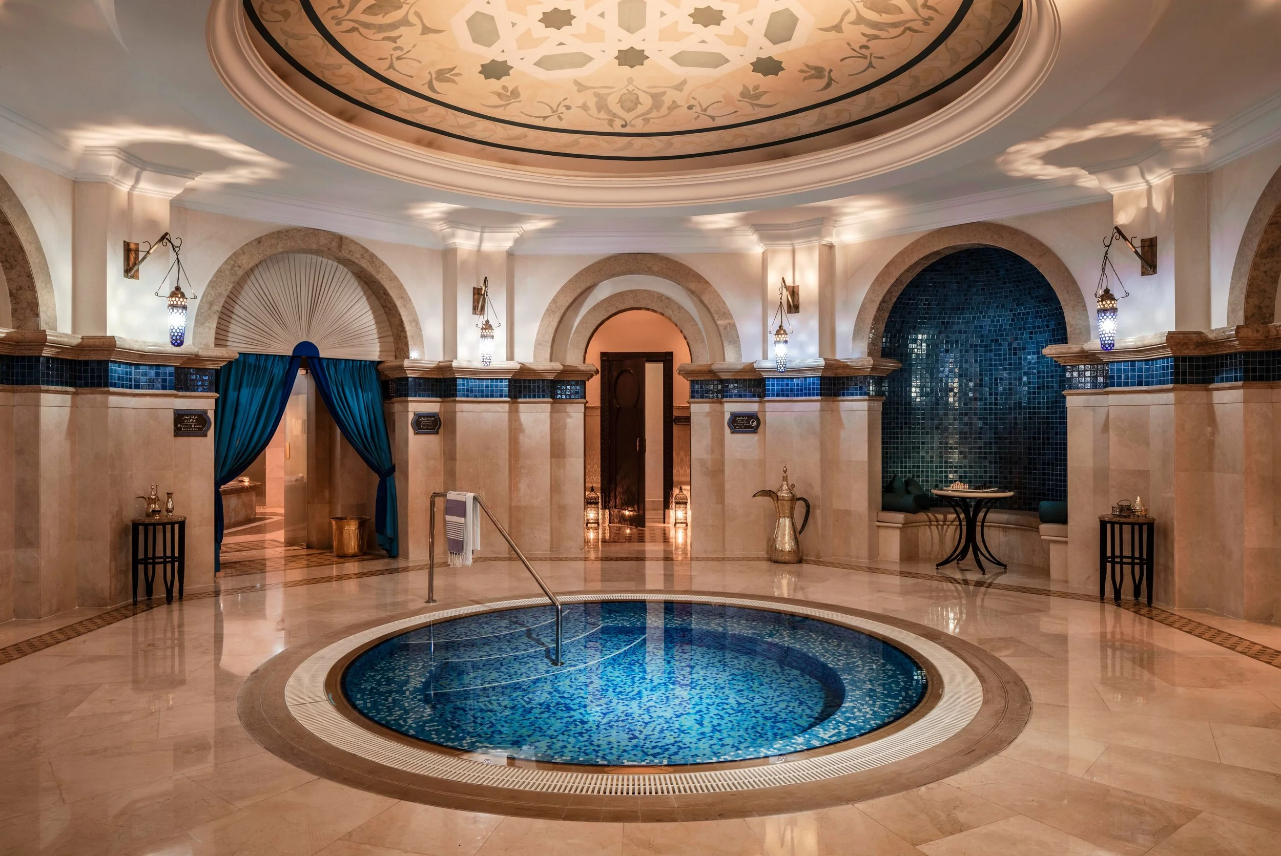 Luxury hotel lobby with ornate ceiling, blue mosaic fountain in the center, arched doorways, and elegant lighting fixtures.