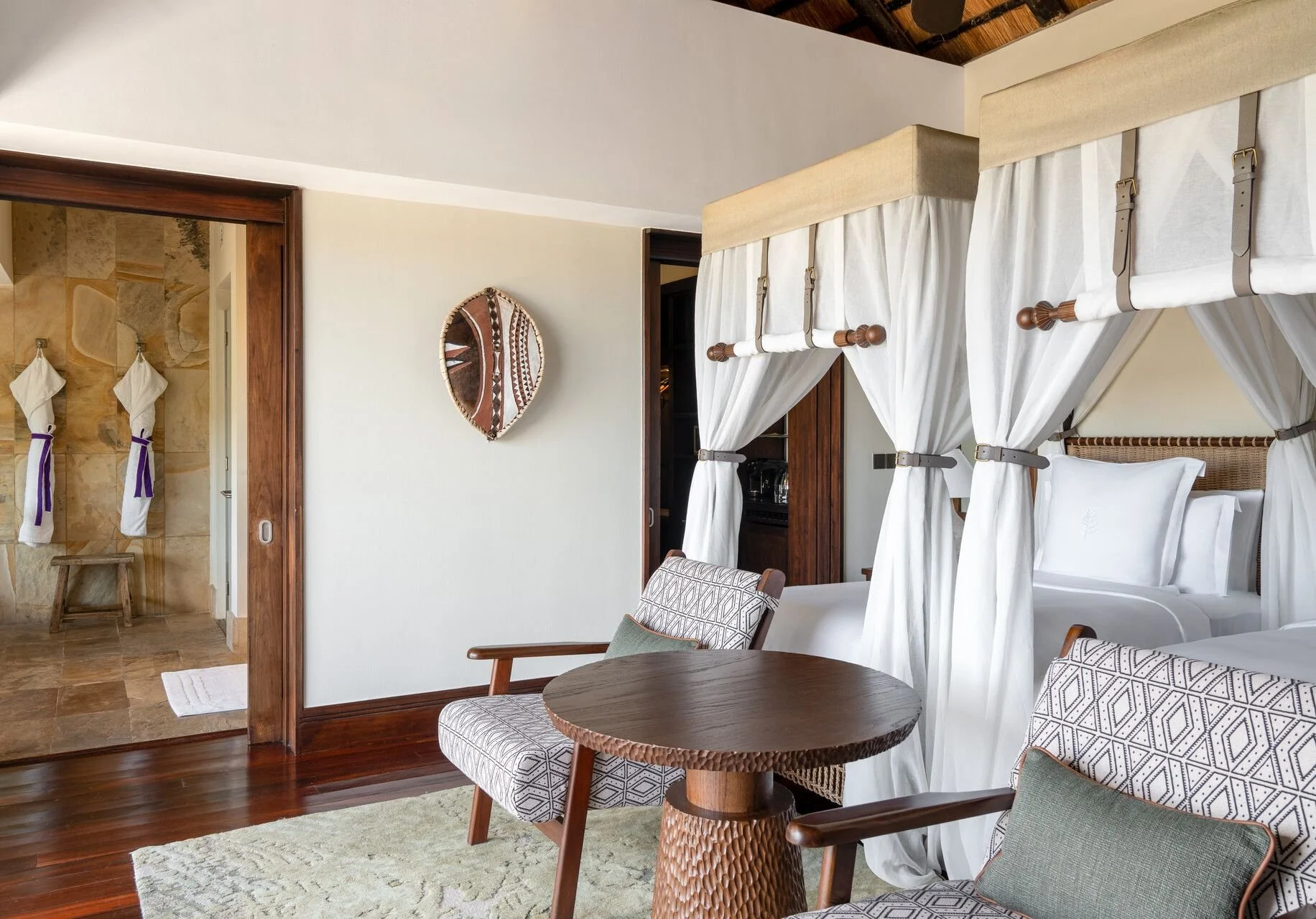 A hotel room with a white four-poster bed with curtains, wooden furniture, chairs, and a wall mirror.