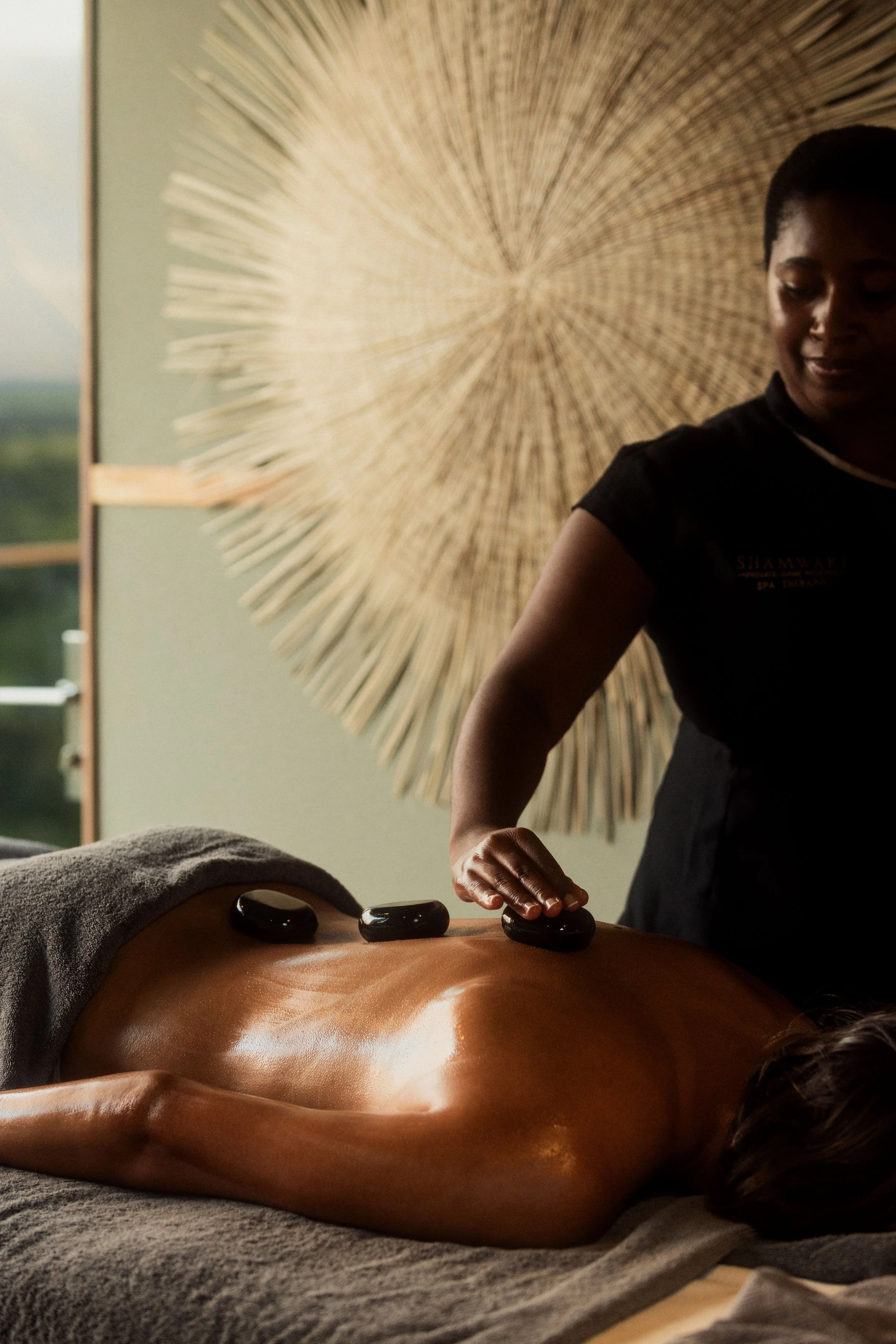 A woman lying face down on a massage table with three stones placed on her back, receiving a hot stone massage. A therapist is placing one of the stones on her back, and a decorative woven wall hanging is visible in the background.