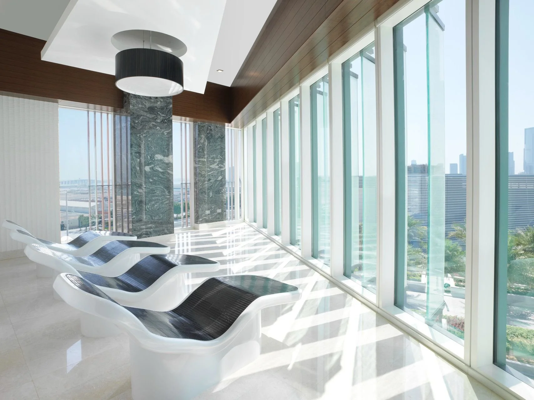 Modern lounge area with white and black lounge chairs, large floor-to-ceiling windows, view of skyscrapers, and a ceiling with a black and white hanging light fixture.