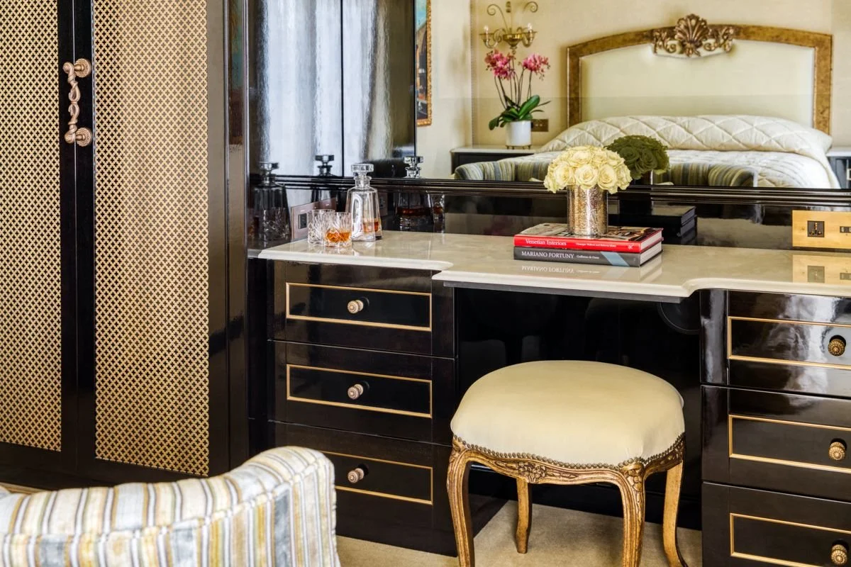 A hotel room with a black painted dresser, a bar tray with glasses and a decanter, a mirror, a bed with floral bedding, a potted plant, a mirror, and decorative wall lamps.