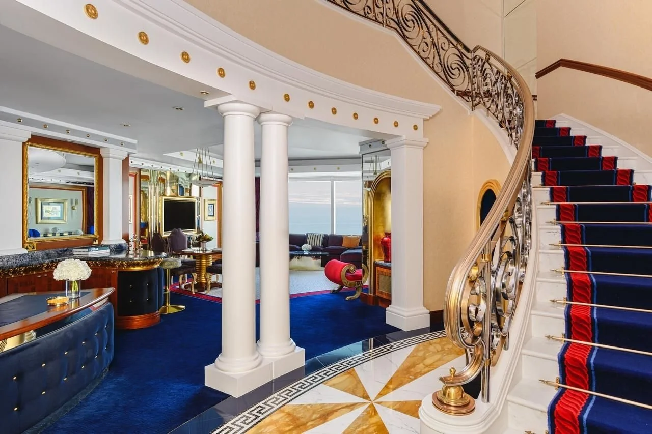 Luxurious hotel interior with a grand staircase covered in navy blue and red carpet, a sitting area with a large window overlooking the ocean, and elegant decor including gold accents and columns.