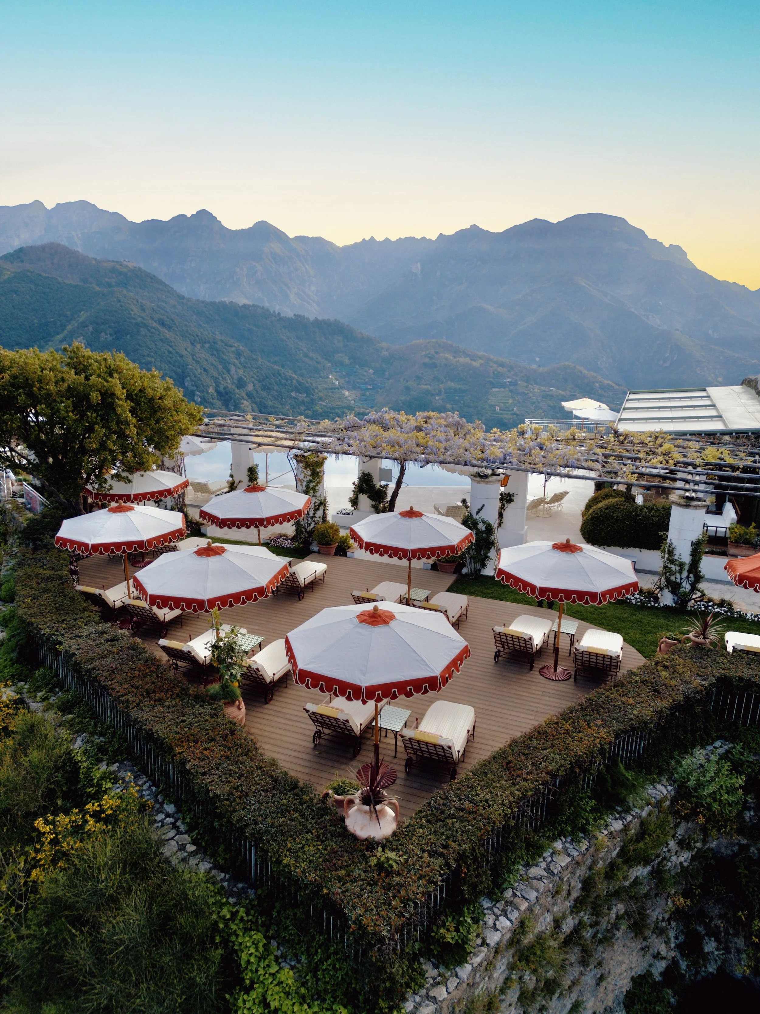 Outdoor terrace with white and red umbrellas, lounge chairs, and lush greenery against a mountainous landscape at sunset.