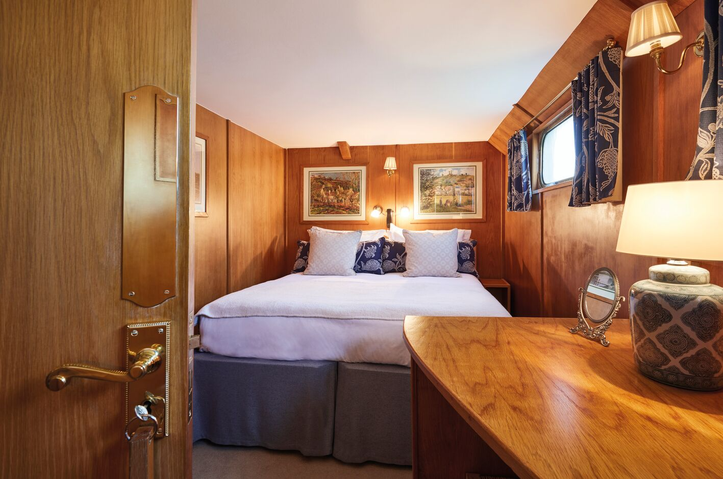 Interior of a cozy bedroom with wooden walls, a large bed with white and patterned pillows, framed landscape paintings above the bed, small wall-mounted lamps, curtains matching the pillows, a bedside table, and a wooden dresser with a table lamp and a small mirror.