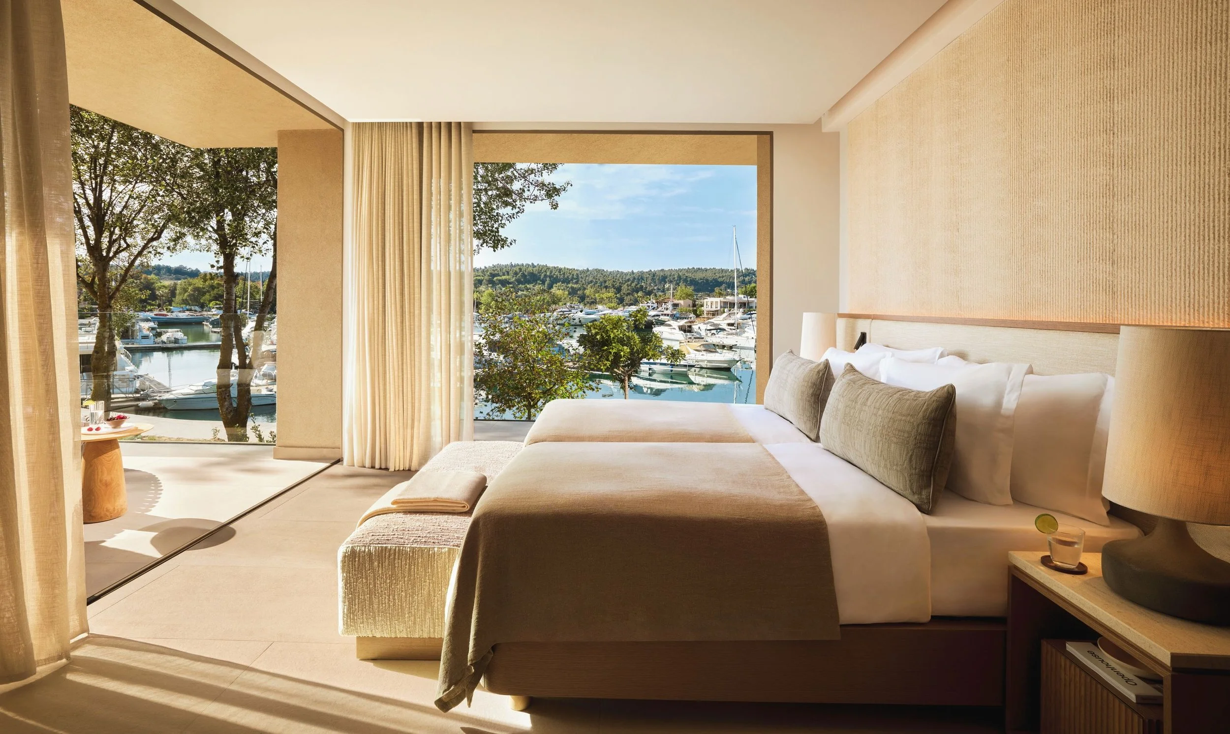 A modern hotel room with a large window view of a marina filled with boats and surrounded by trees. The room has a bed with beige and green pillows covered in beige bedding, beige curtains, and a bedside table with a lamp.