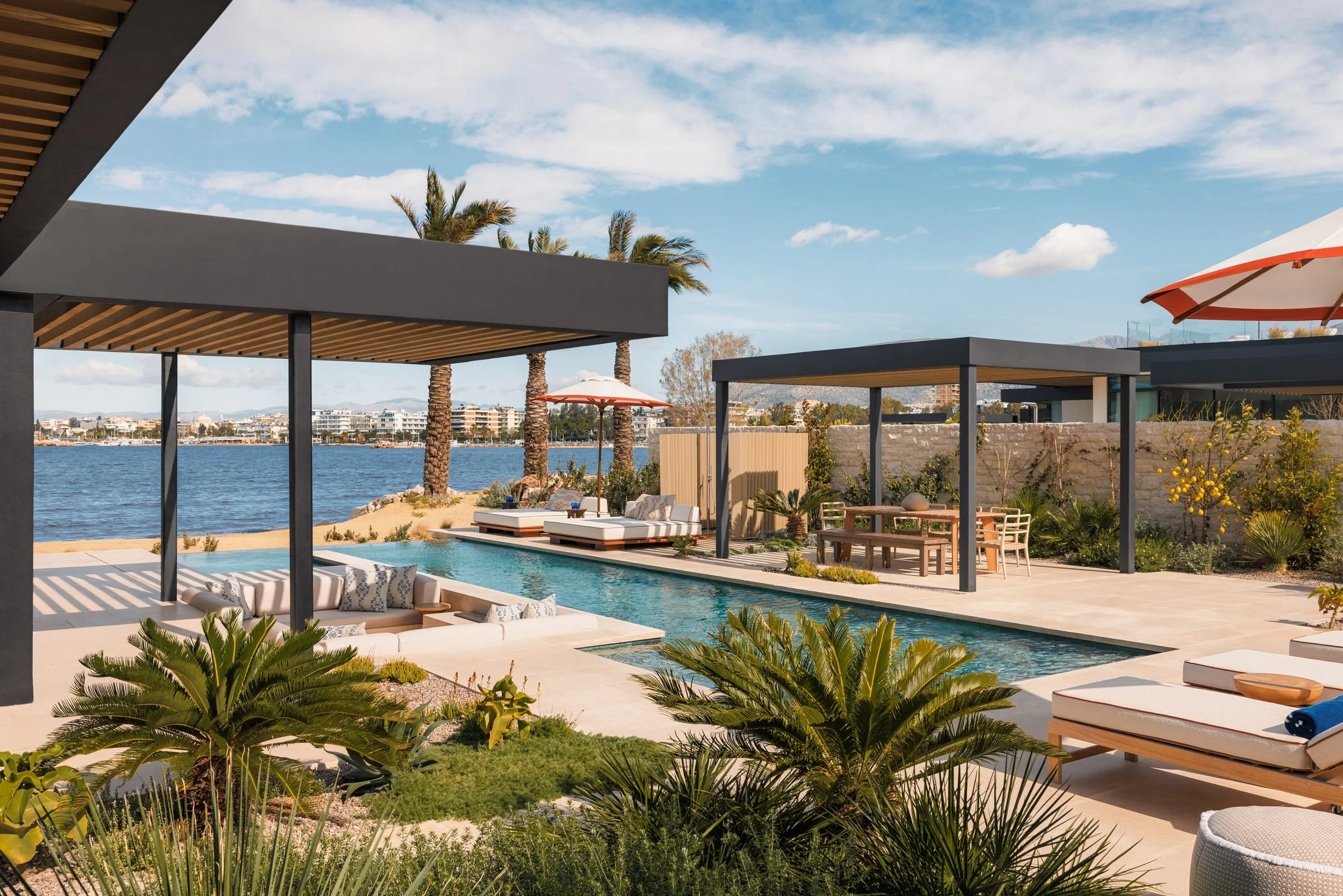 Luxury outdoor pool area with lounge chairs, shaded seating, lush greenery, and palm trees overlooking a body of water under a partly cloudy sky.