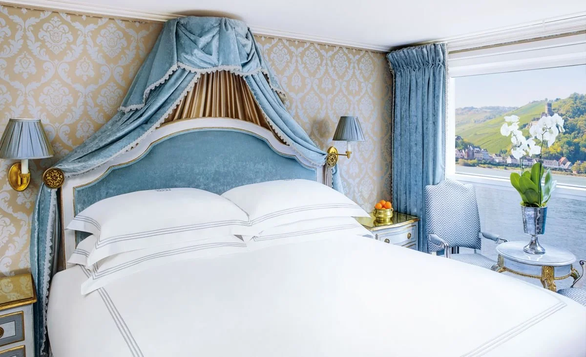 Luxurious hotel bedroom with a vintage-style bed, blue velvet headboard, gold accents, and patterned wallpaper. The room has a large window with blue curtains, showing a scenic view of green hills and a river. A side table with a flower pot and a bowl of fruit, and a blue-and-white patterned chair are visible.