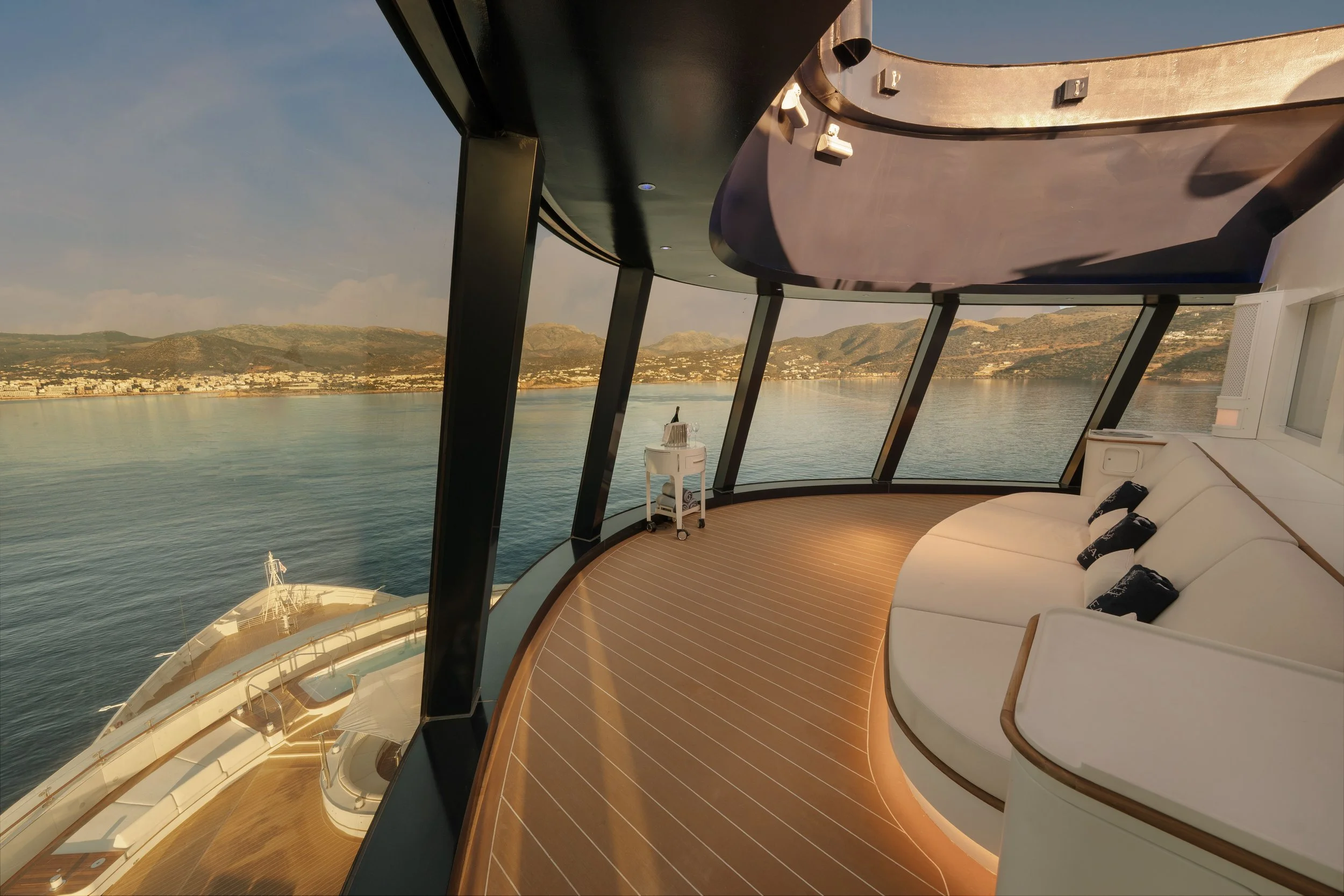 Interior of a modern cruise ship with a curved glass wall, overlooking calm blue water and distant land with hills. There is a white couch with pillows, a small table, and polished wooden flooring.