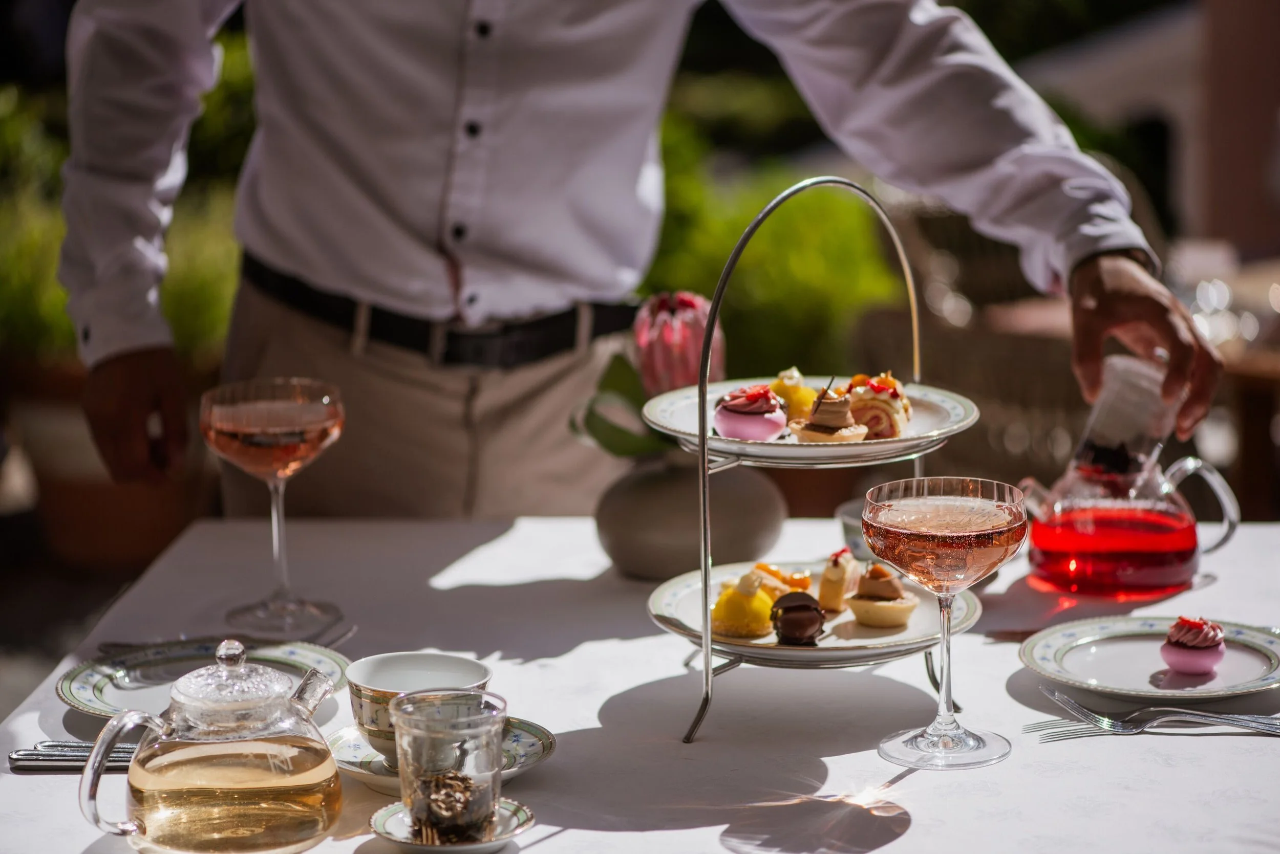 A person in a white shirt serves desserts on a table set outdoors. The table features a tiered stand with assorted pastries and small cakes, surrounded by teacups, glasses of pink liquid, and a teapot.