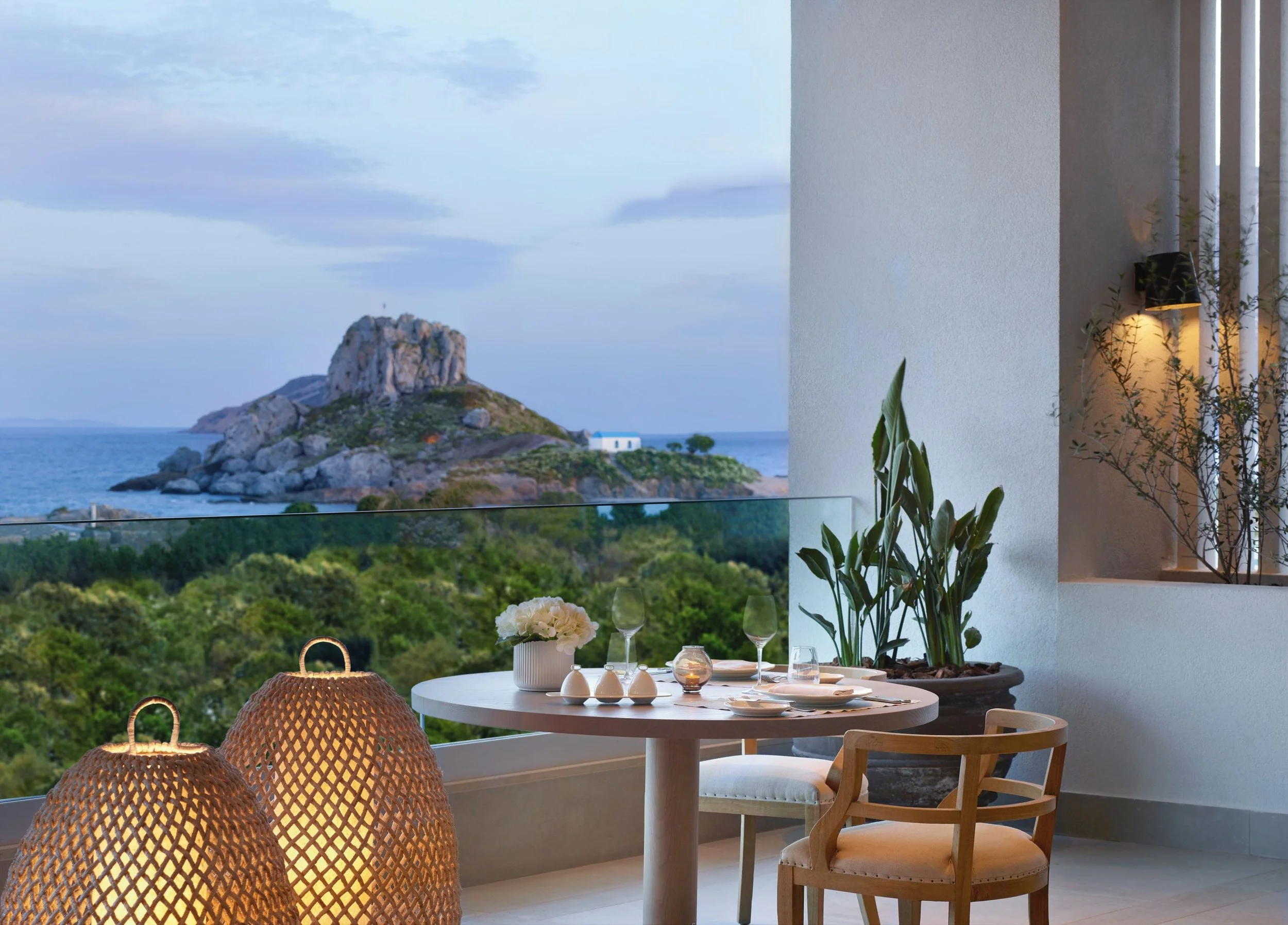 A balcony dining area overlooking a scenic ocean view with a rocky island and greenery. The table is set with glassware, a white flower arrangement, and candles, with woven lanterns hanging nearby. Indoor plants and contemporary furniture are visible inside.