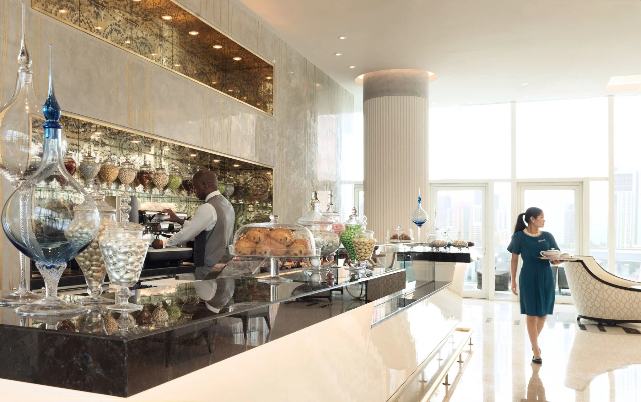 A modern, elegant cafe or pastry shop interior with large windows, showcasing a counter with baked goods, candies in glass jars, and a barista preparing drinks. Two women are present, one working behind the counter and another walking with a tray of plates.