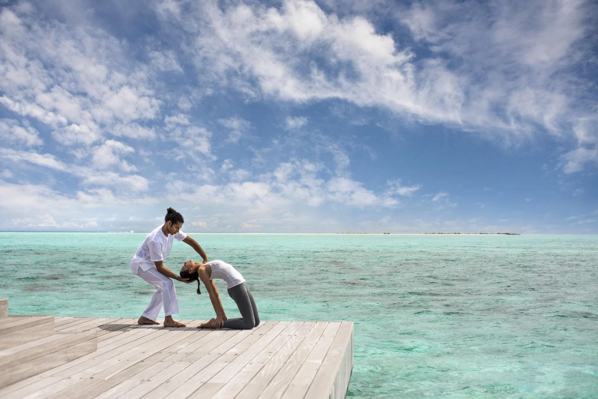 Two people practicing yoga on a wooden dock by the ocean, with a woman in a headstand position and a man supporting her, under a partly cloudy sky.