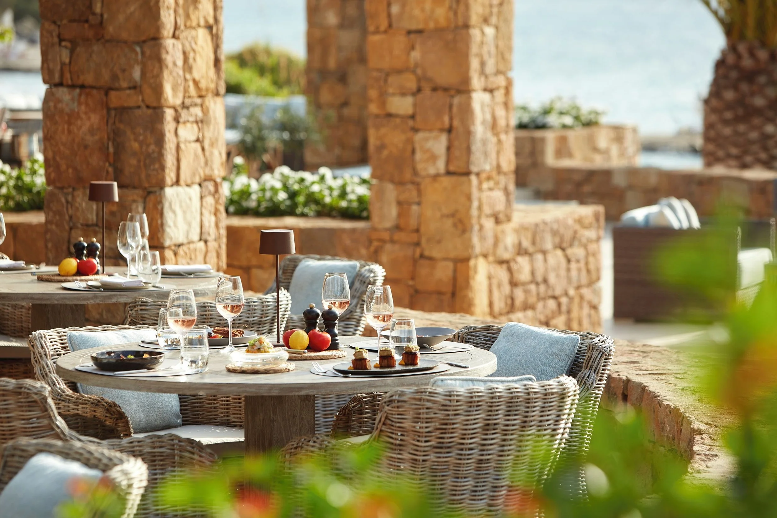 Outdoor restaurant setup with tables and chairs, set for dining with wine glasses, plates, and silverware, near a rustic brick structure overlooking water.