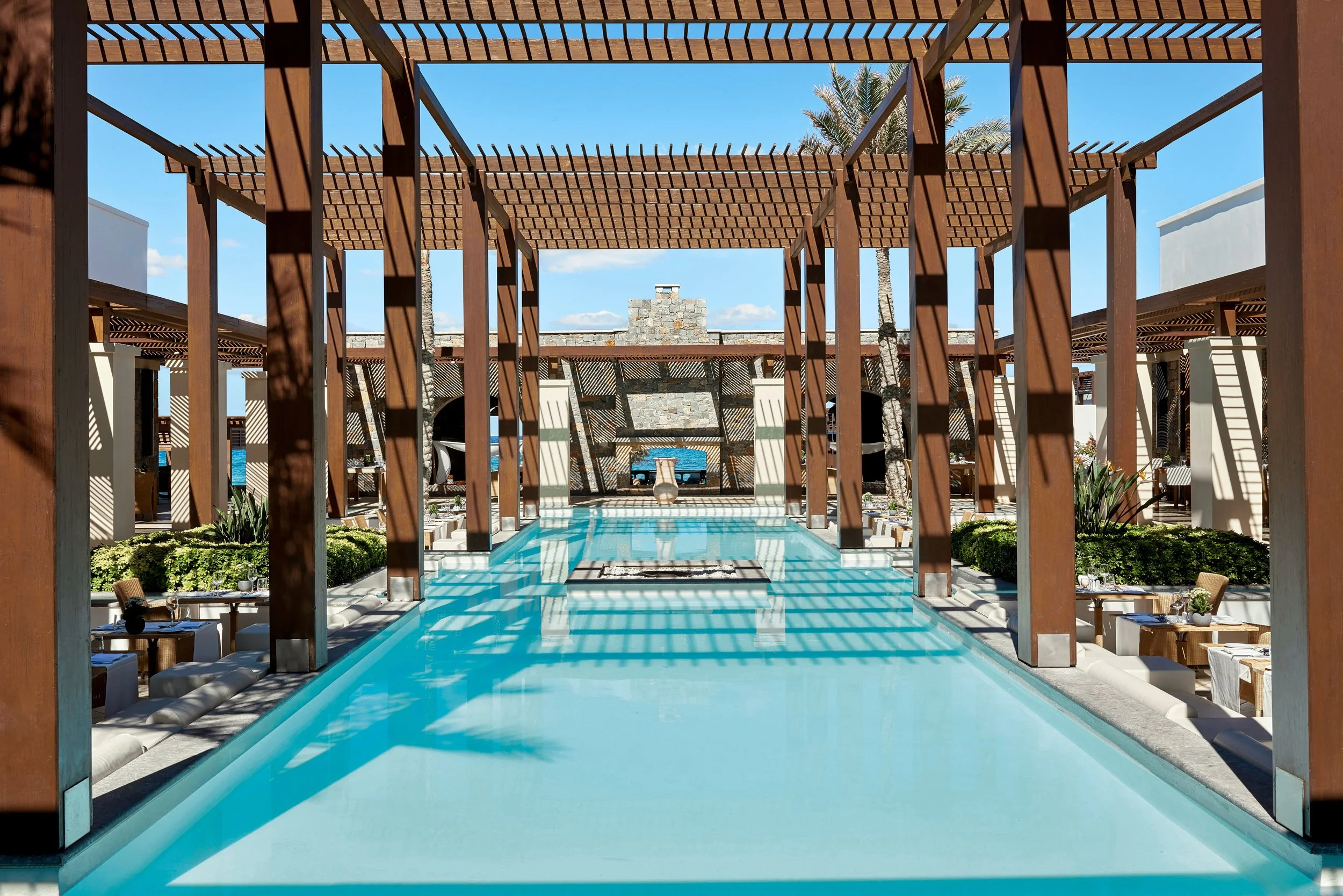A swimming pool in a resort with wooden pergolas providing shade, surrounded by lounge chairs, with palm trees and a stone fireplace in the background under a clear blue sky.