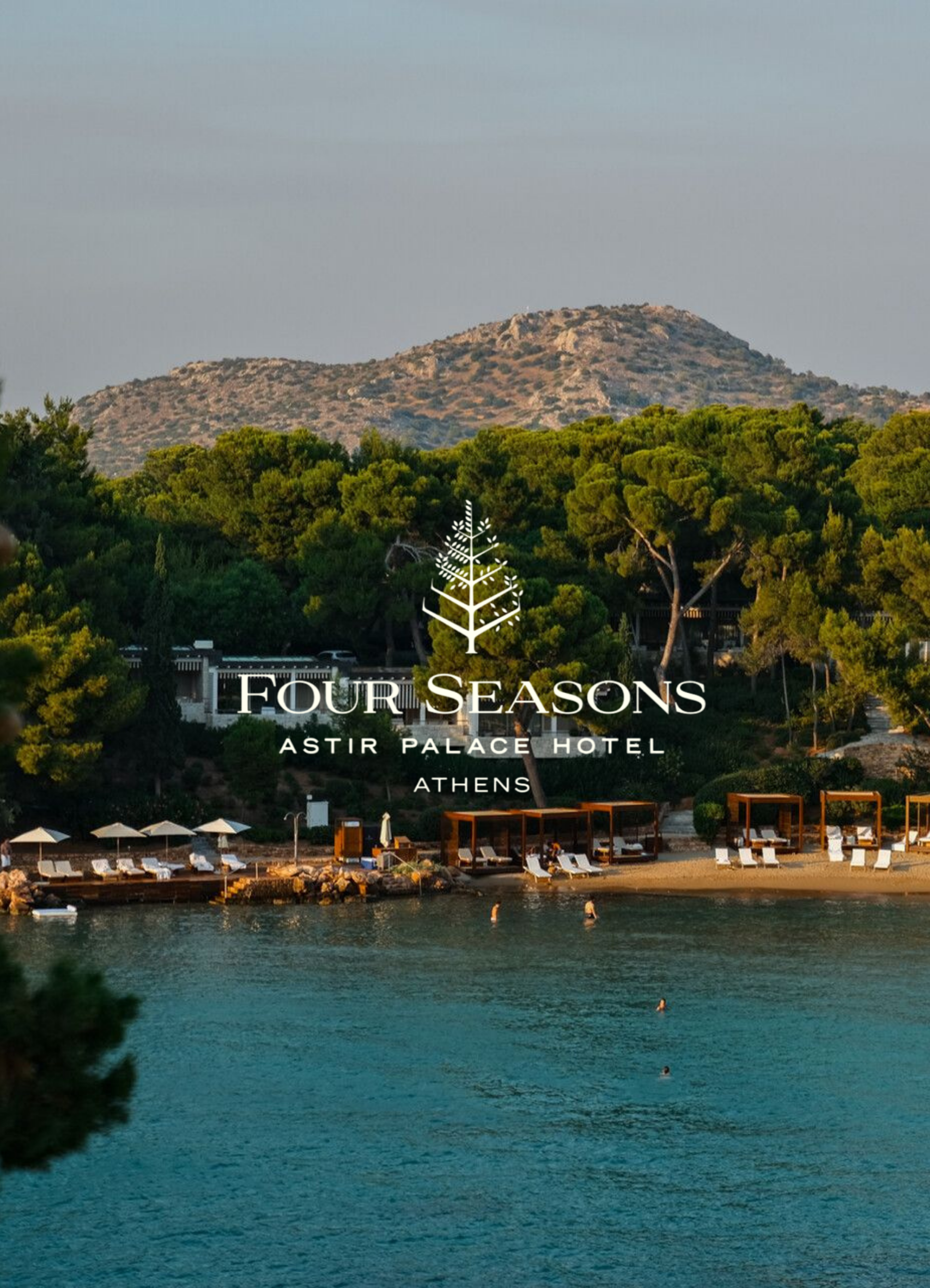 A scenic view of a beach with lounge chairs and umbrellas, calm water with a few swimmers, lush green trees, and a rocky hill in the background under a clear sky.