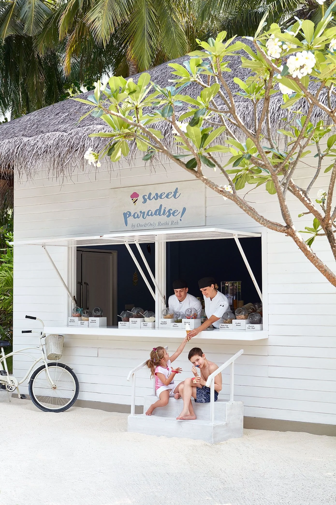 A small ice cream stand called 'Sweet Paradise' with two employees serving ice cream. In front, two children sitting on a white bench passing or receiving ice cream, with a girl handing a cone to a boy. There is a white bicycle parked nearby, and palm trees can be seen in the background.