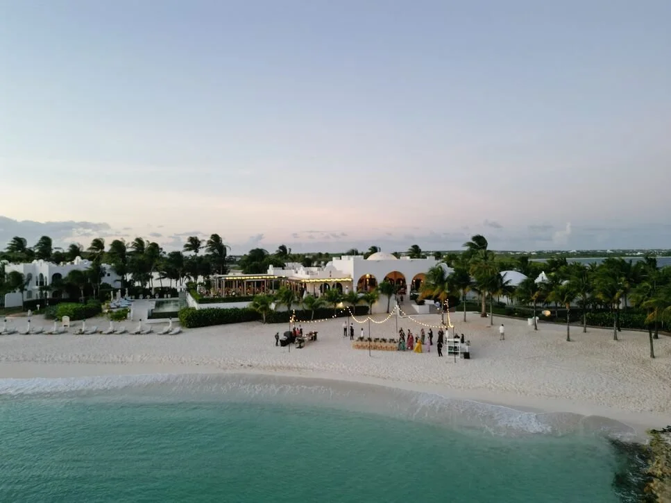 Beachside venue with white sand, palm trees, and a beachfront building decorated with string lights, set against the ocean with a clear sky.