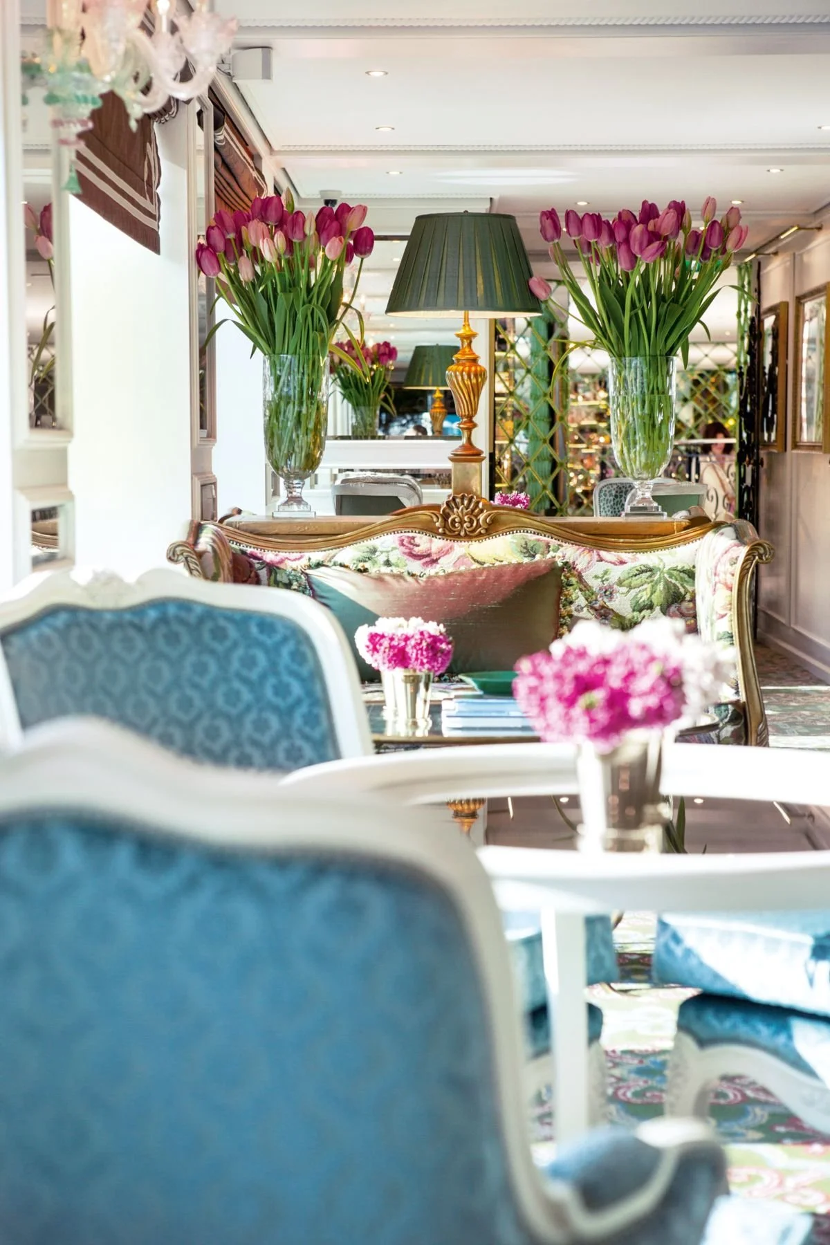 Elegant living room with blue and floral upholstered chairs, a floral sofa, and glass vases with pink tulips on tables. Two table lamps with green shades are on a side table. The room has decorative mirrors, framed artwork, and a patterned carpet.