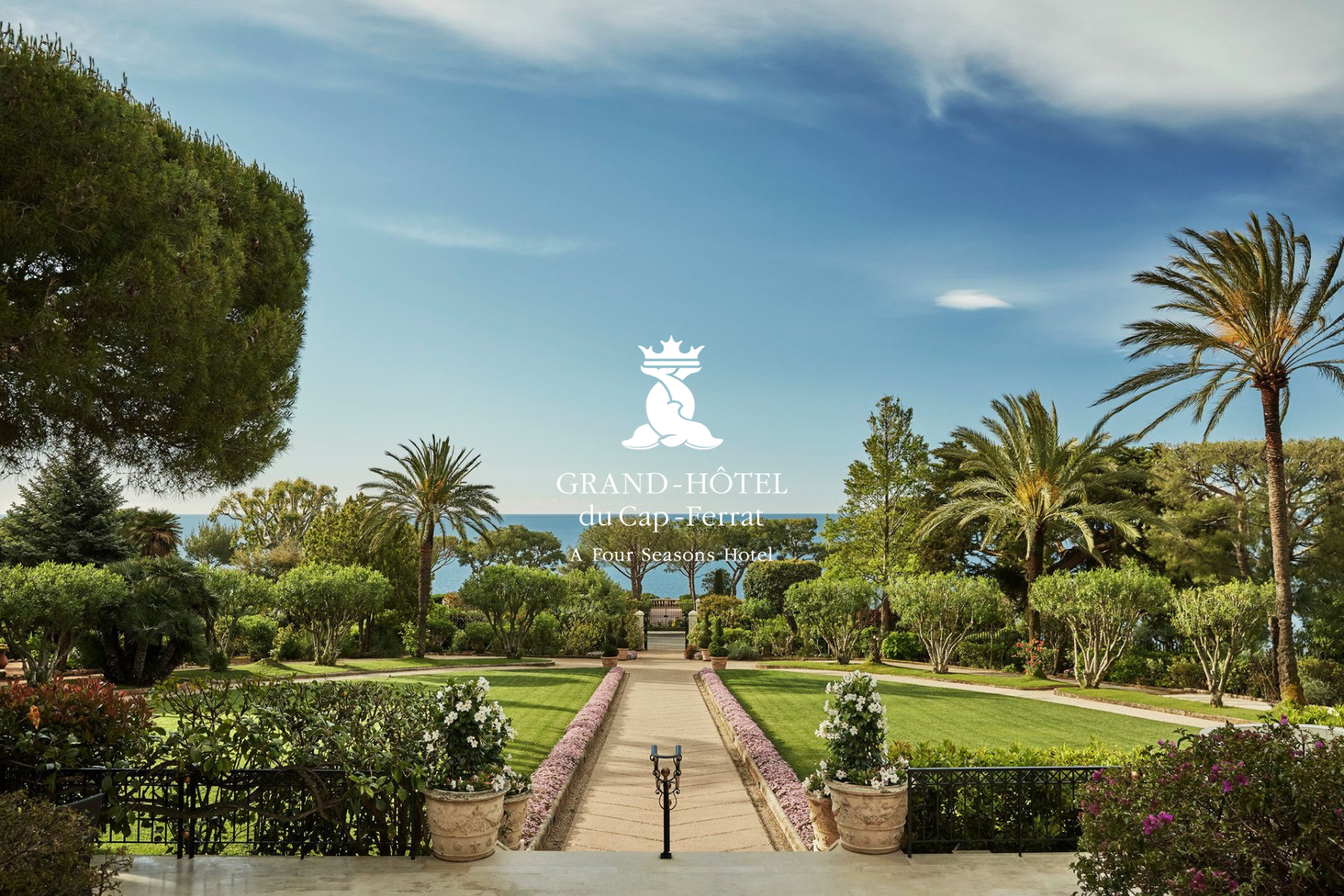 A lush green garden with palm trees and flowering bushes leads to a distant view of the sea under a partly cloudy sky. The photo features the logo and name of the Grand-Hôtel du Cap-Ferrat, a Four Seasons Hotel, superimposed in the center.