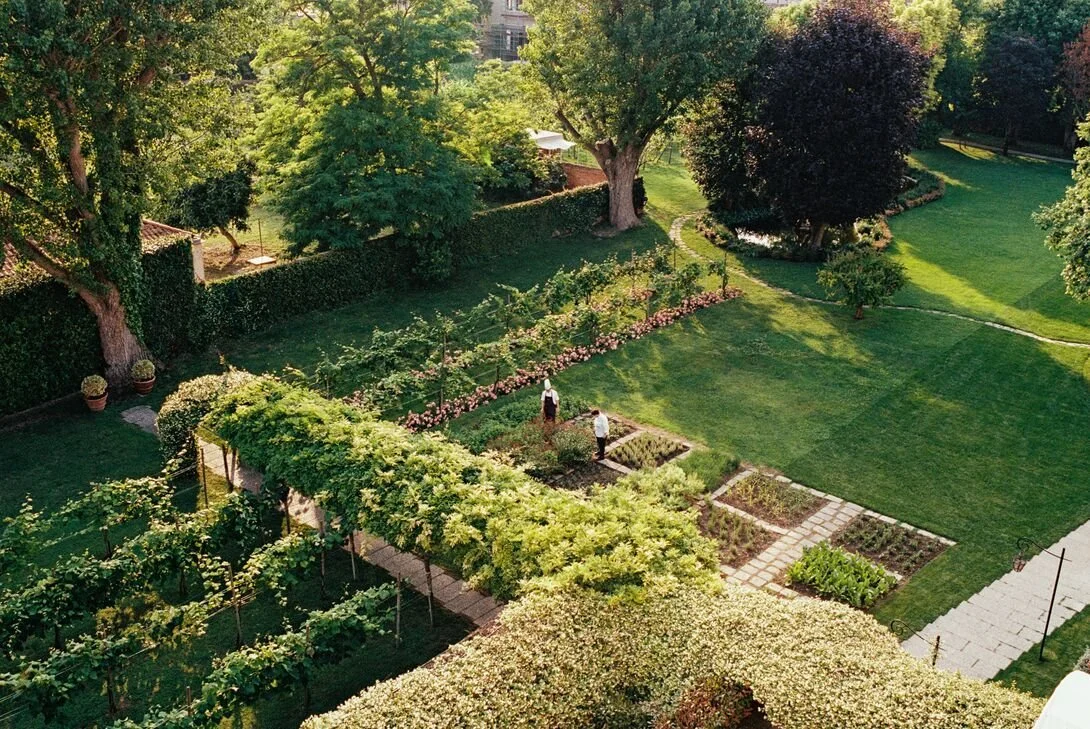 A lush, green garden with trees, shrubbery, and flower beds, and two people tending to the plants.