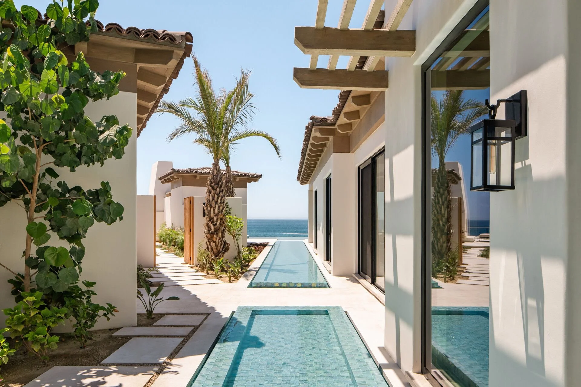 View of a modern house with floor-to-ceiling windows, a narrow pool, and tropical plants, with the ocean and clear blue sky in the background.